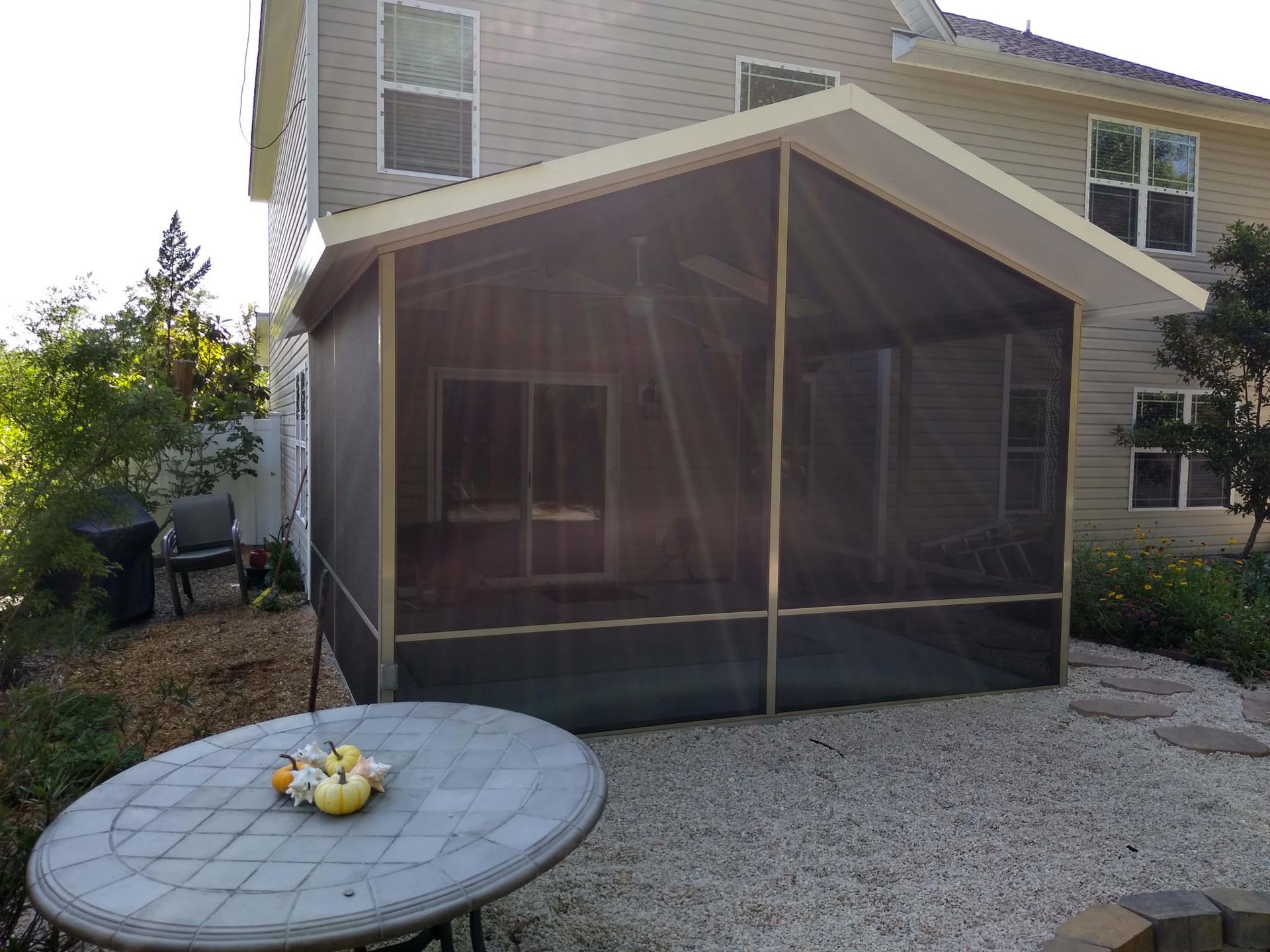A screened-in patio attached to the back of a beige house with a gravel yard and a round tiled table in the foreground.