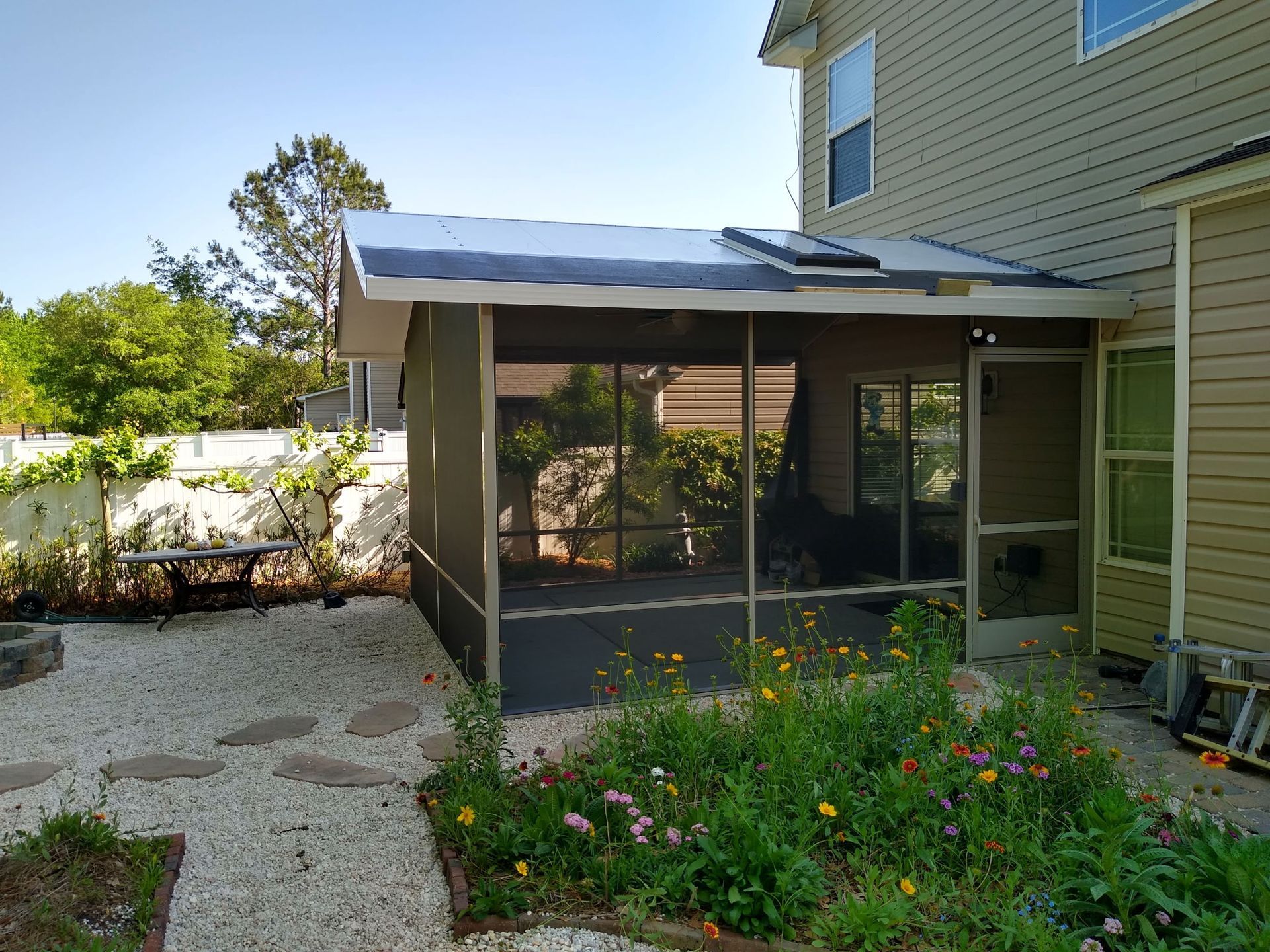A screened-in patio attached to a two-story tan house, featuring a light-colored roof, garden, and gravel yard.