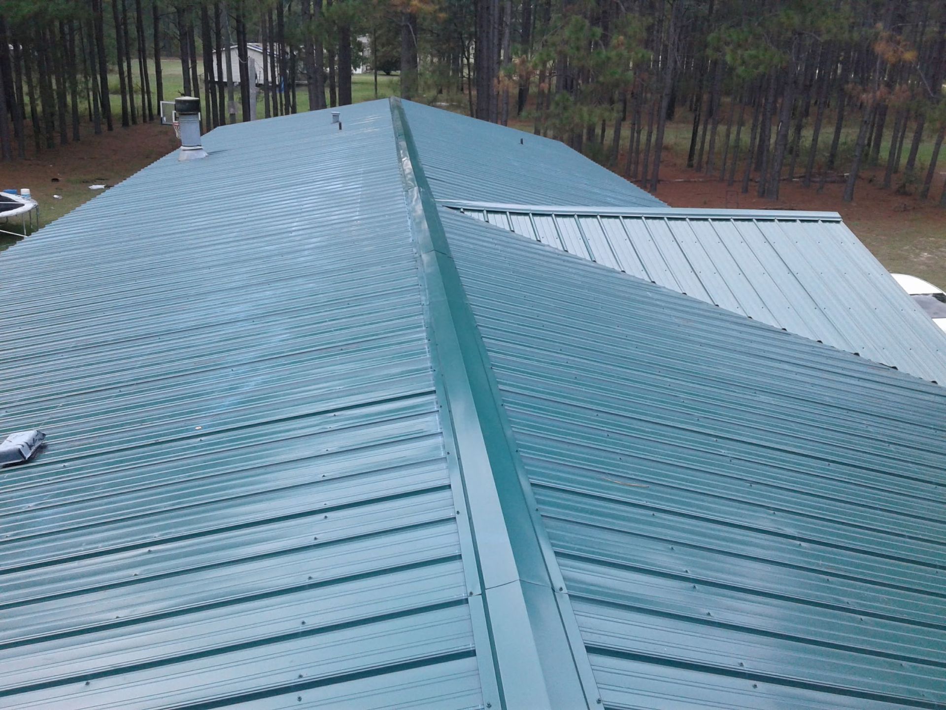 An elevated view of a green metal roof with a central ridge cap and a side section, set against a backdrop of trees.