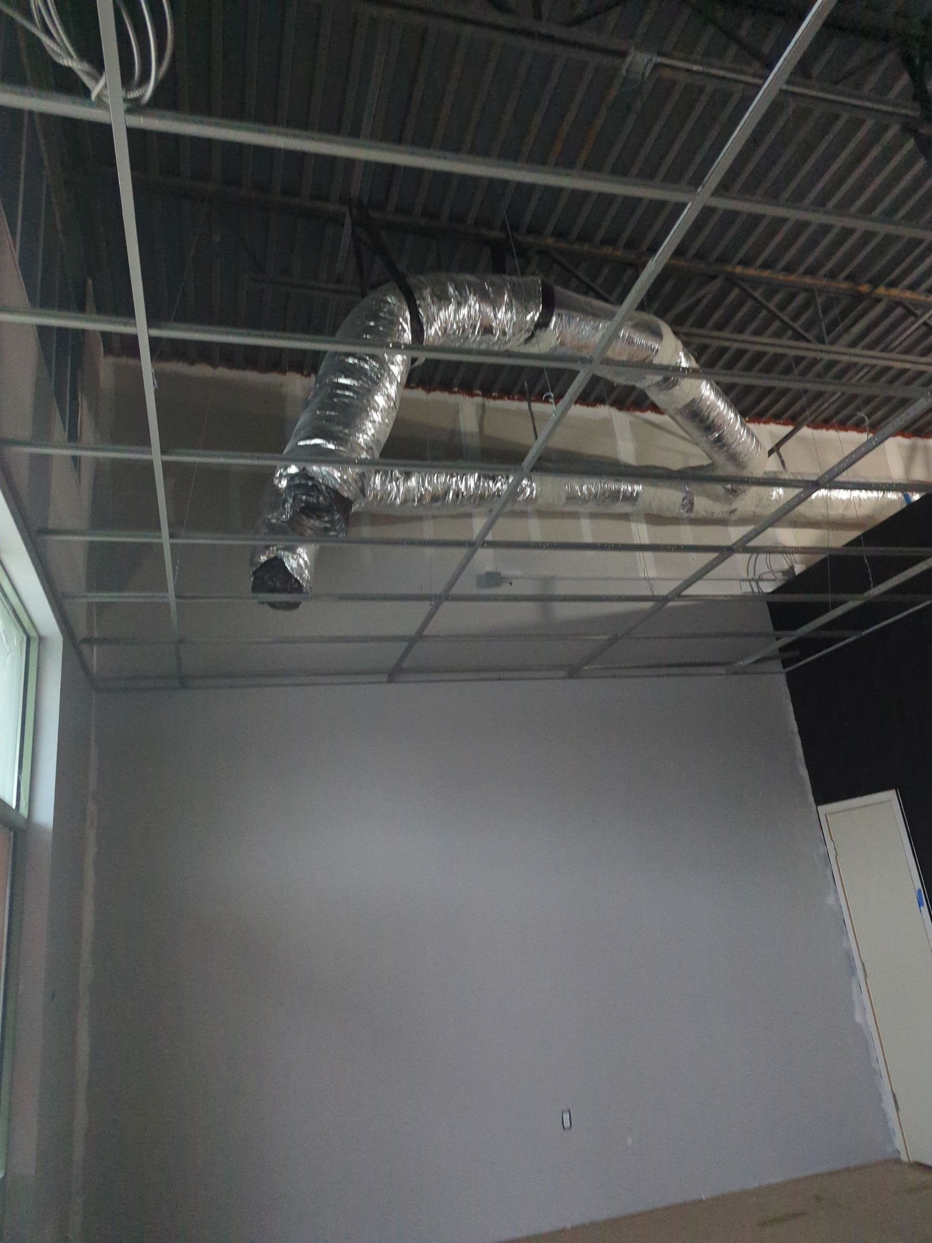 An unfinished room with a suspended metal grid ceiling and exposed silver ductwork hanging above gray walls.