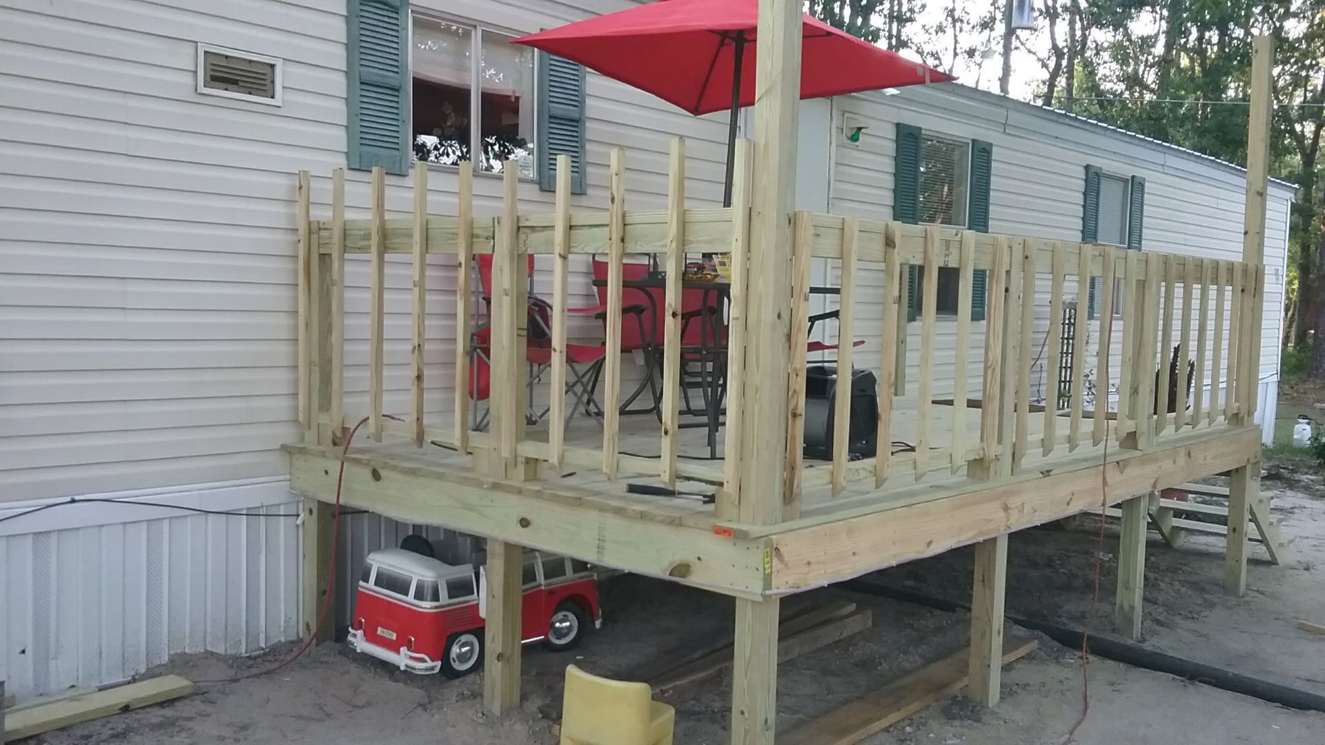 A wooden deck attached to a light-colored mobile home, featuring a red umbrella, outdoor chairs, and a toy fire truck below.