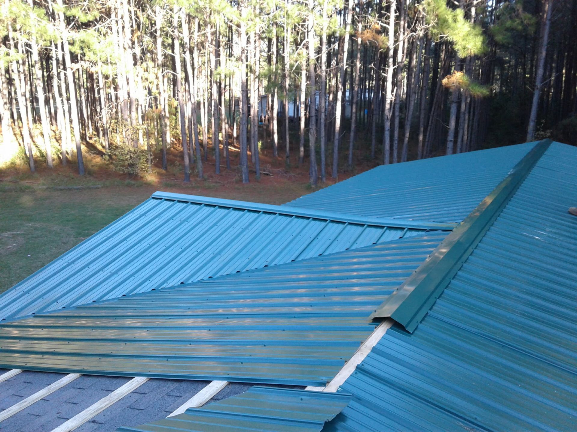 An unfinished green metal roof with exposed underlayment, viewed from above with a forest in the background.
