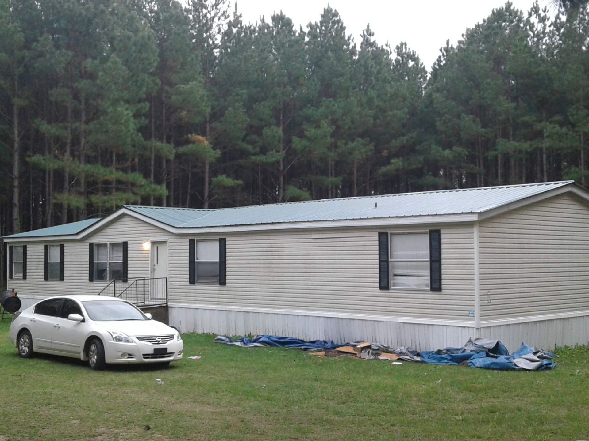 A single-story, light-colored mobile home sits in a grassy yard in front of a dense pine forest with a white sedan parked.