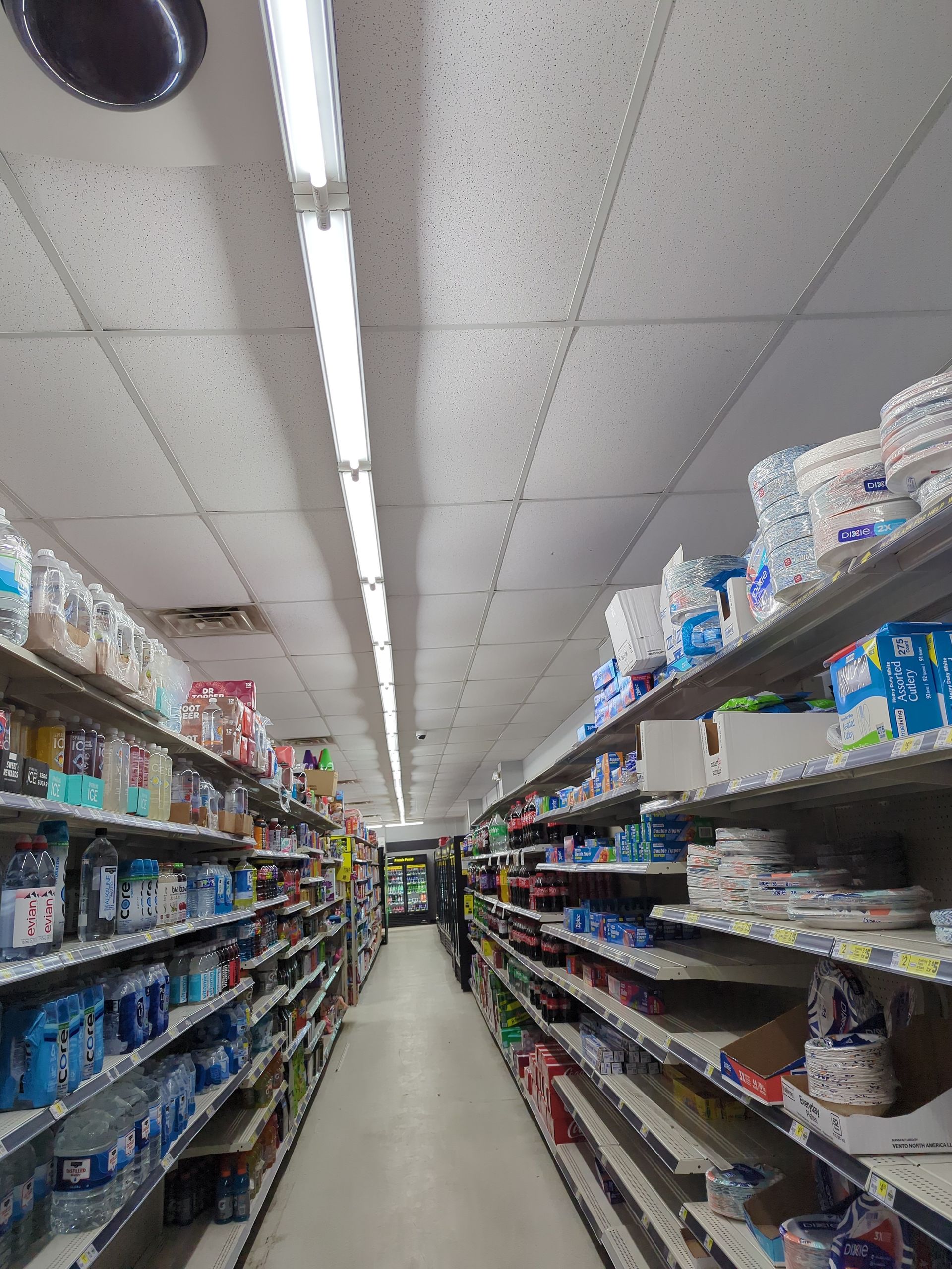 An aisle in a grocery store, lined with shelves filled with various household products and a fluorescent ceiling light.
