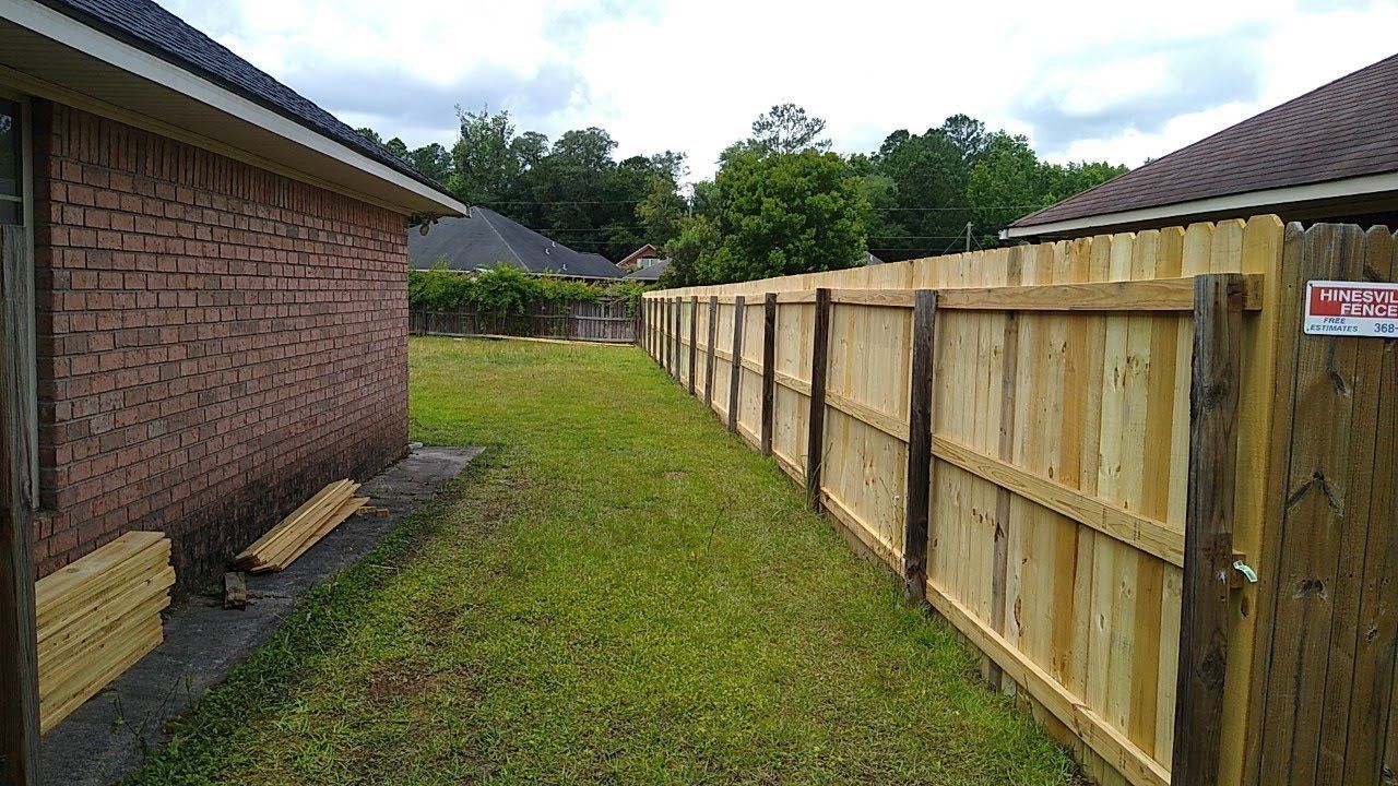 A long, newly constructed wooden fence runs along the side of a brick house, bordering a grassy side yard.