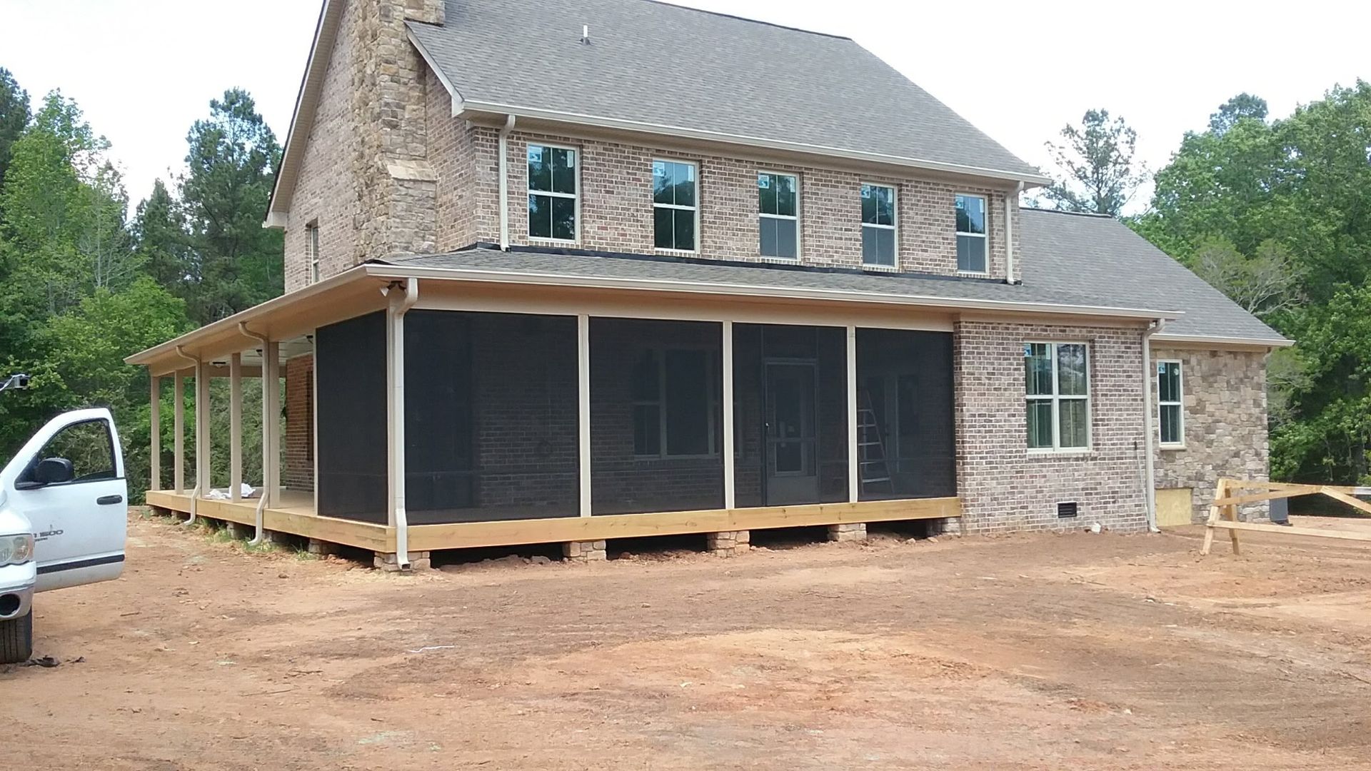 A two-story brick house with a newly constructed, screened-in wooden porch on a dirt lot, featuring a partial white truck.