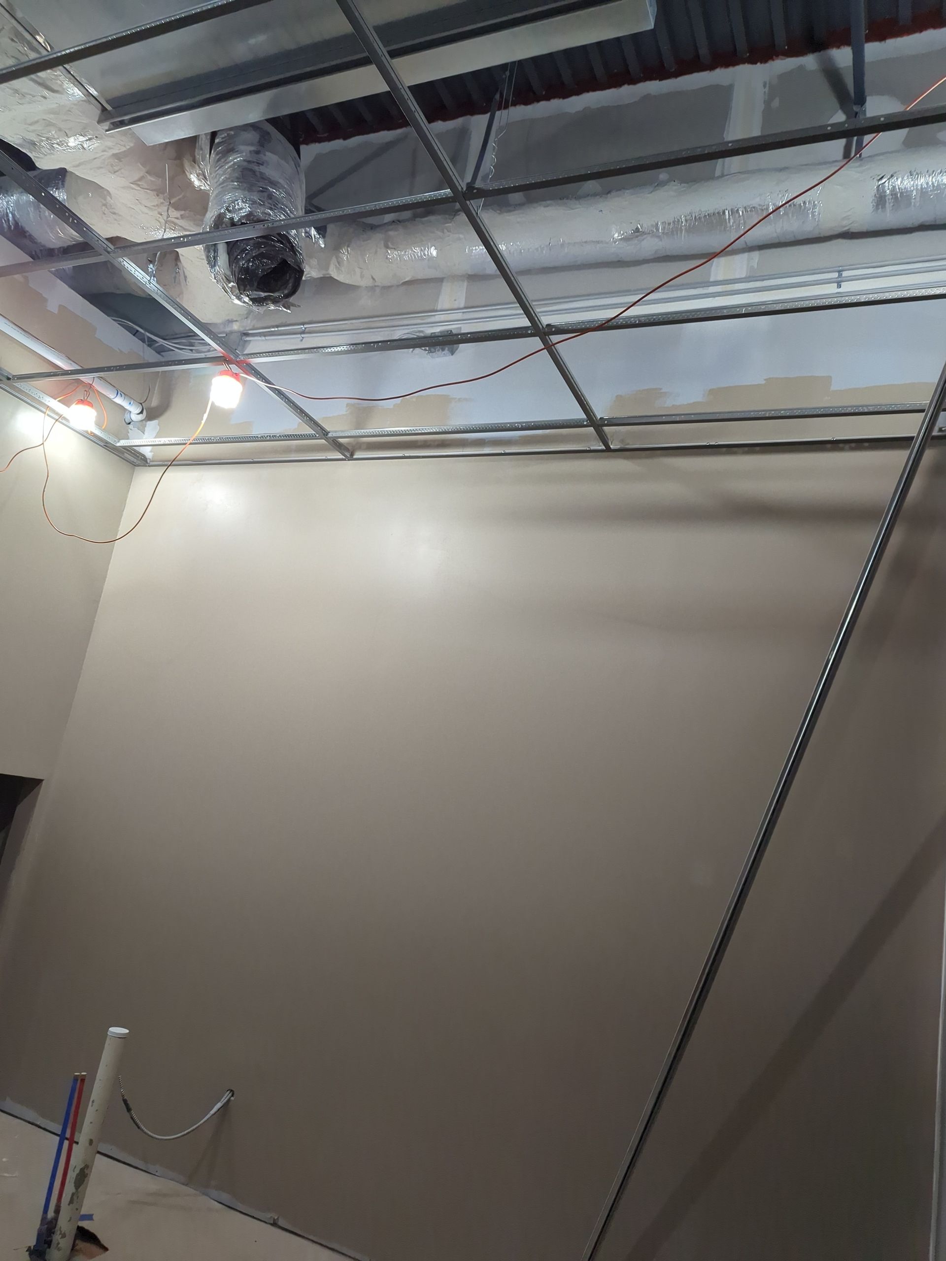 Interior view of a room under construction with a metal ceiling grid and exposed ductwork above a smooth, tan wall.