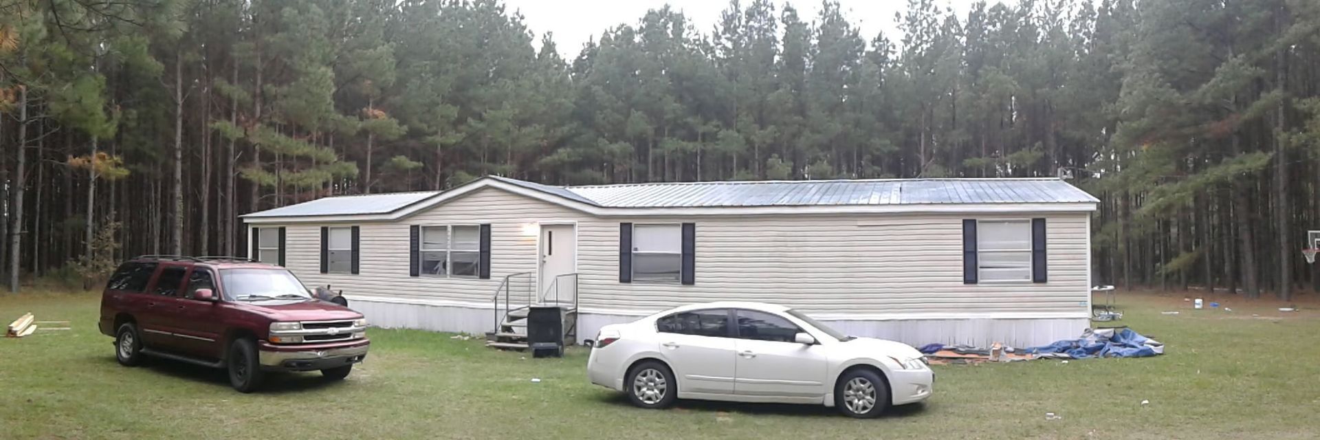 A light-colored mobile home sits on a grassy lawn in front of a dense pine forest, with a maroon SUV and white sedan parked.