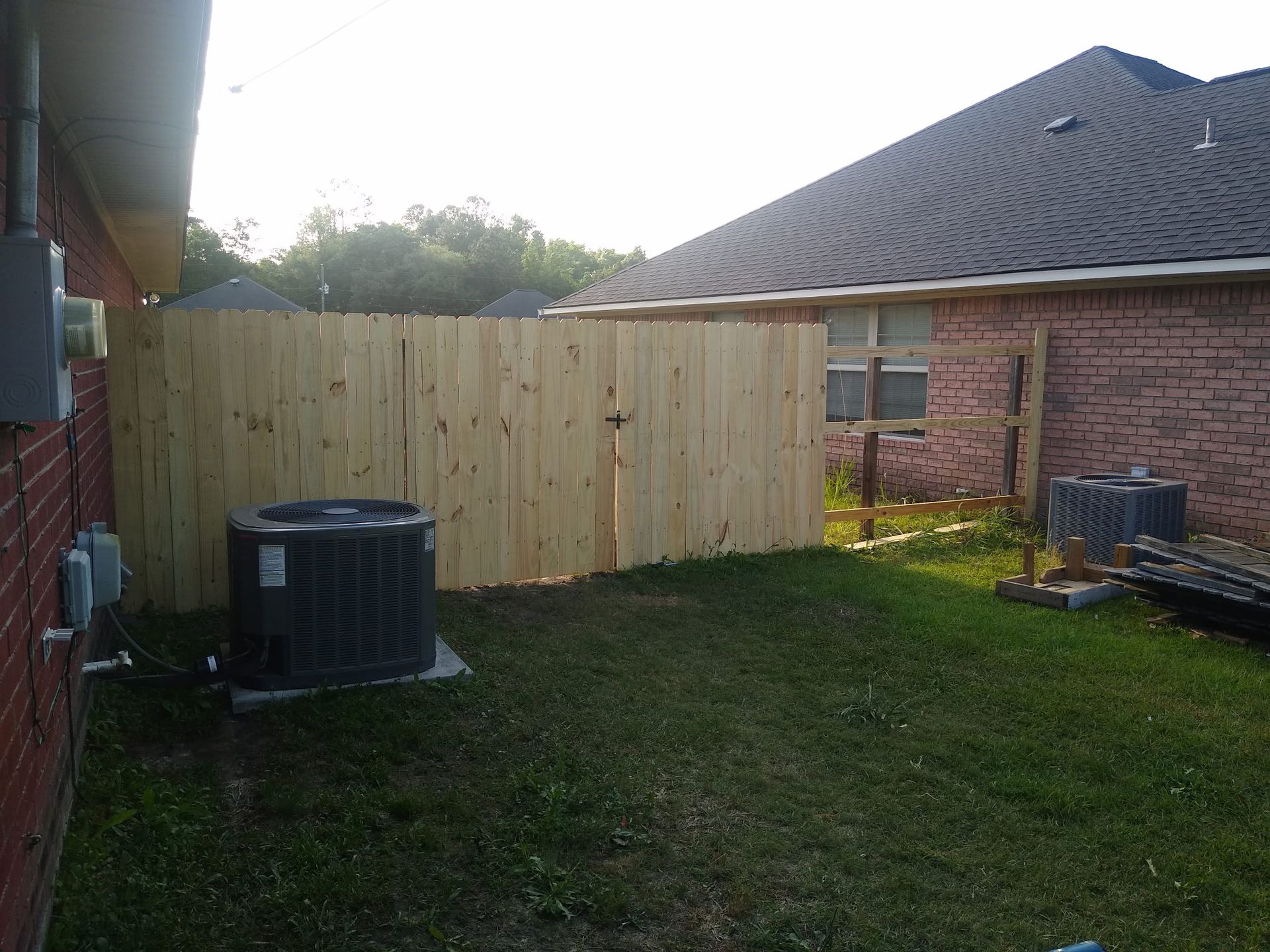 A new wooden privacy fence in a residential backyard, with air conditioning units near brick house walls.