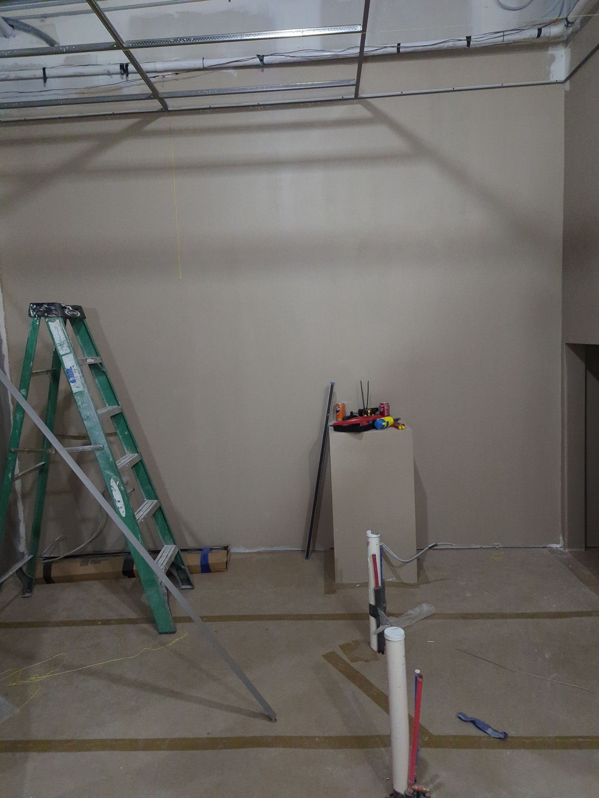 An interior construction site with a green stepladder, a drywall partition, and exposed ceiling grid framing.