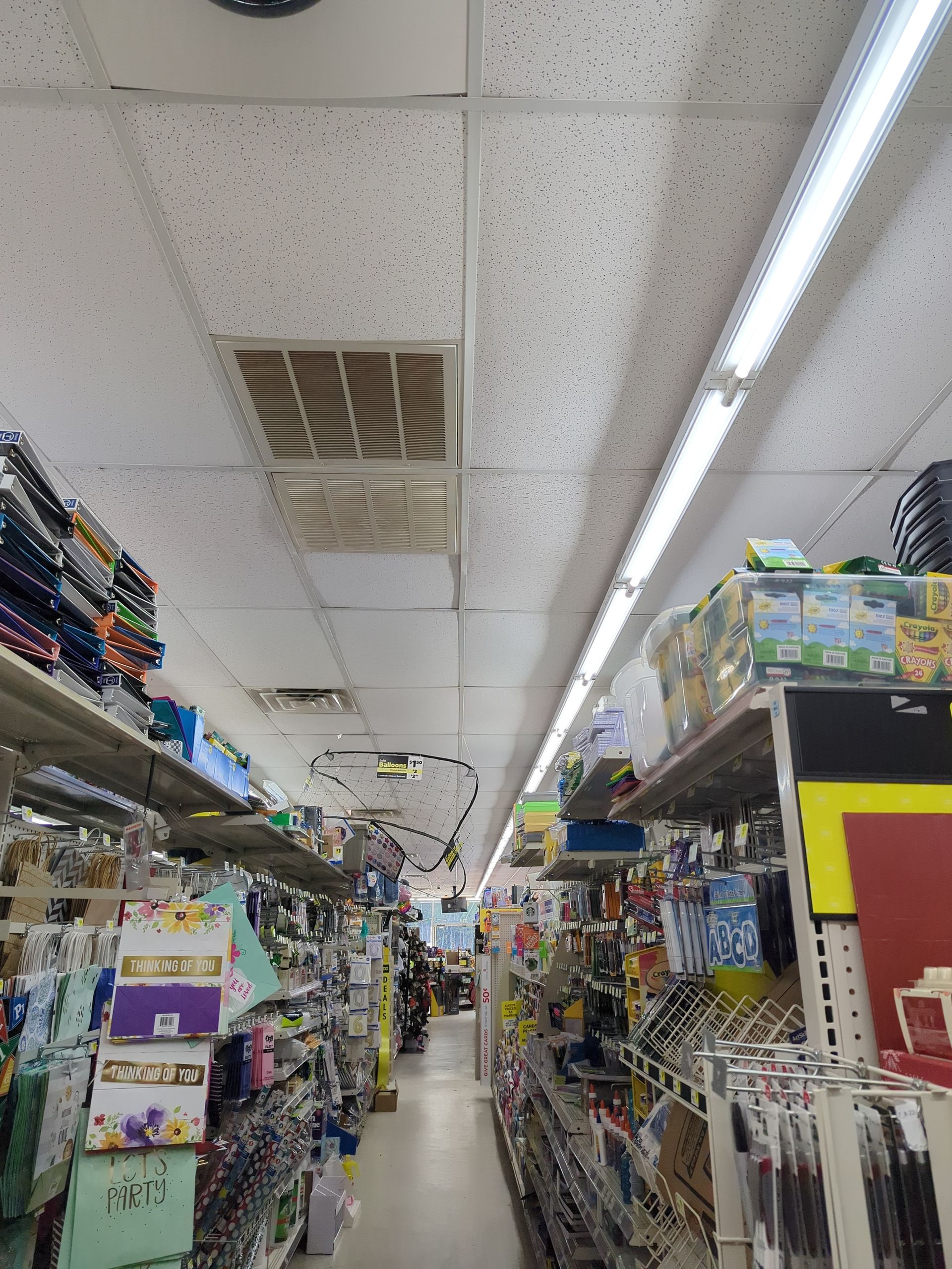 A low-angle view of a store aisle with shelves stocked with various products, a drop ceiling, and bright overhead lighting.