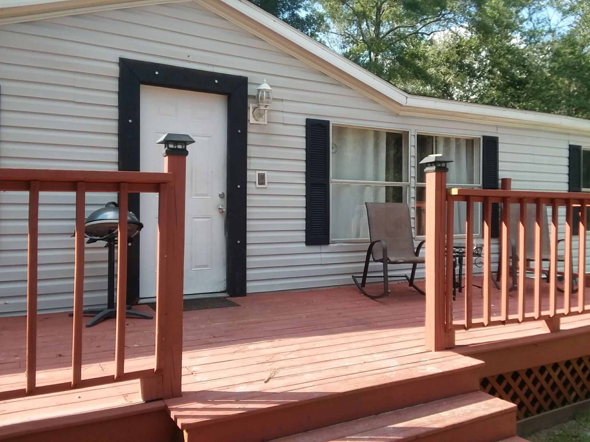 A light gray house with a red deck and railing, featuring a white door, black shutters, and outdoor chairs.