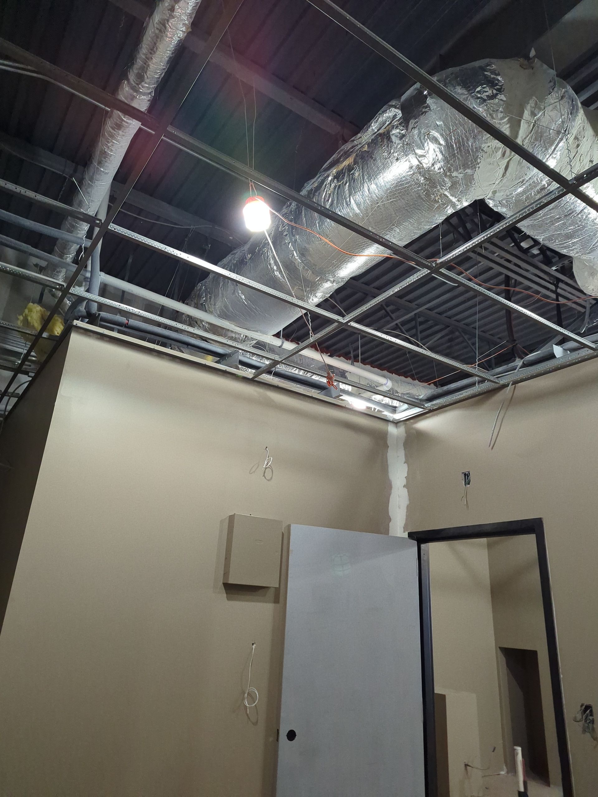 A room under construction with an exposed metal ceiling grid, ductwork, unfinished tan walls, and an open doorway.