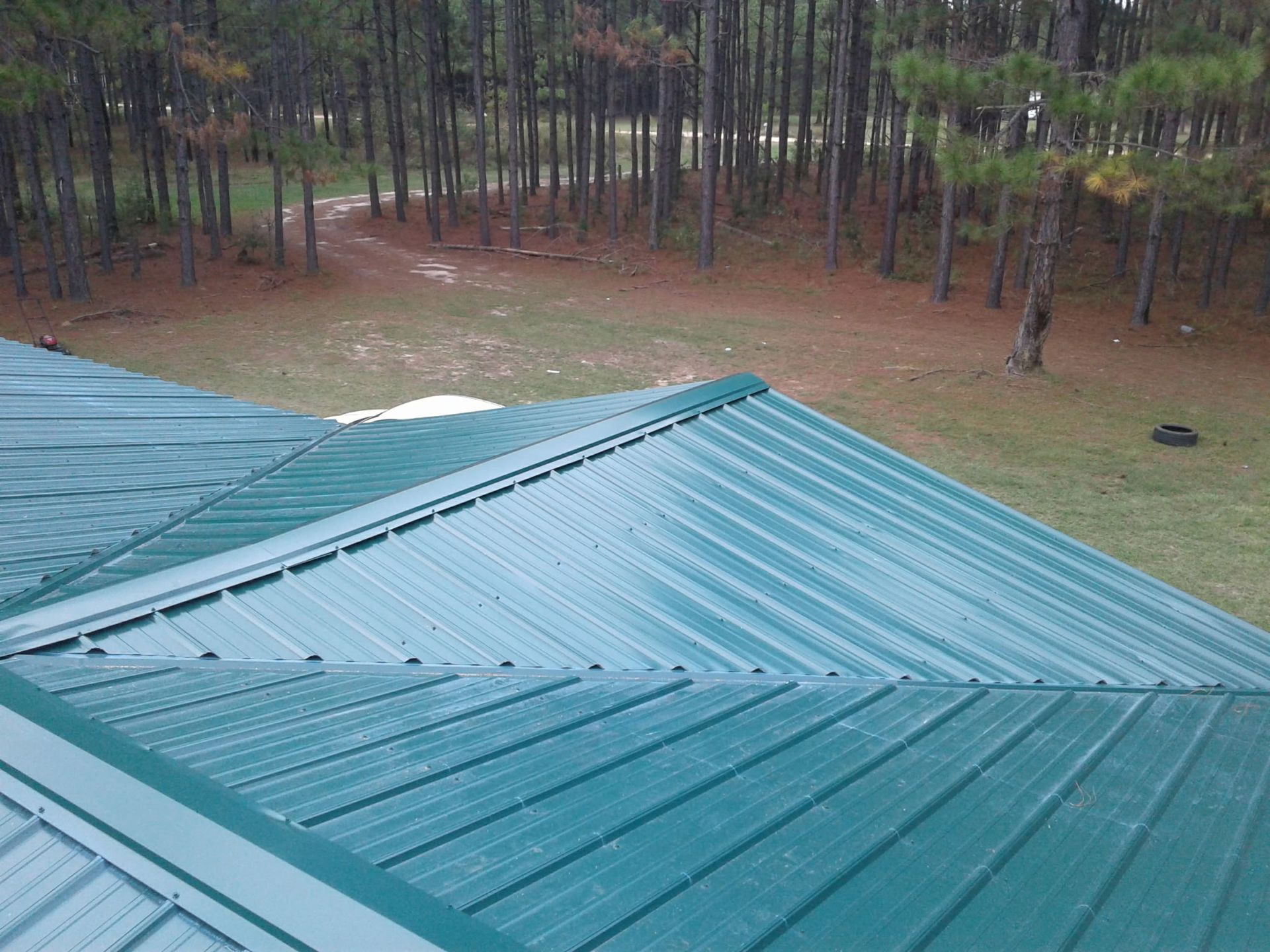 A high-angle view of a green metal roof with panels and ridge caps, overlooking a grassy yard and a dense pine forest.