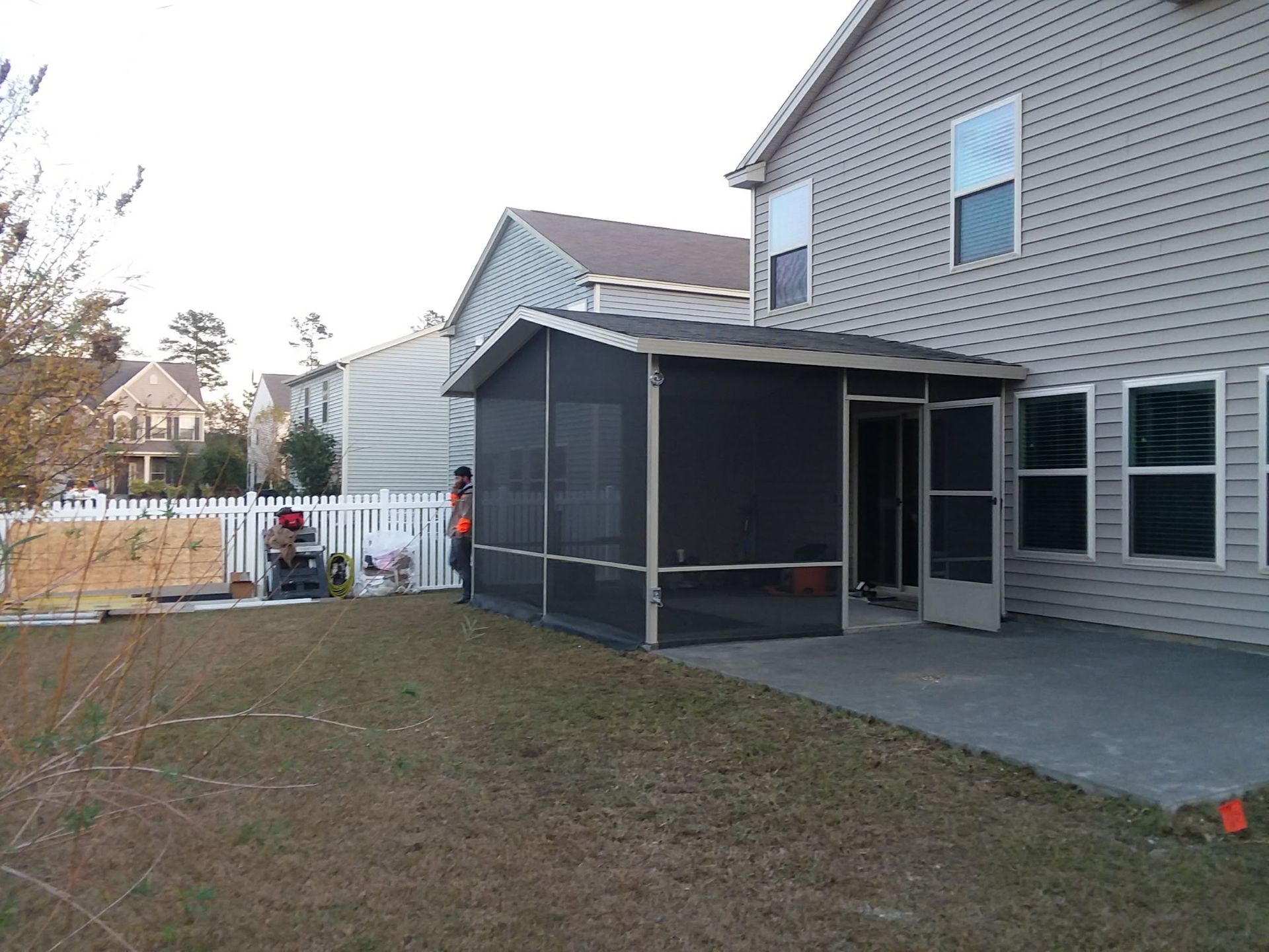 A grey house with a screened-in porch and patio in a suburban backyard, with a person standing by the porch entrance.