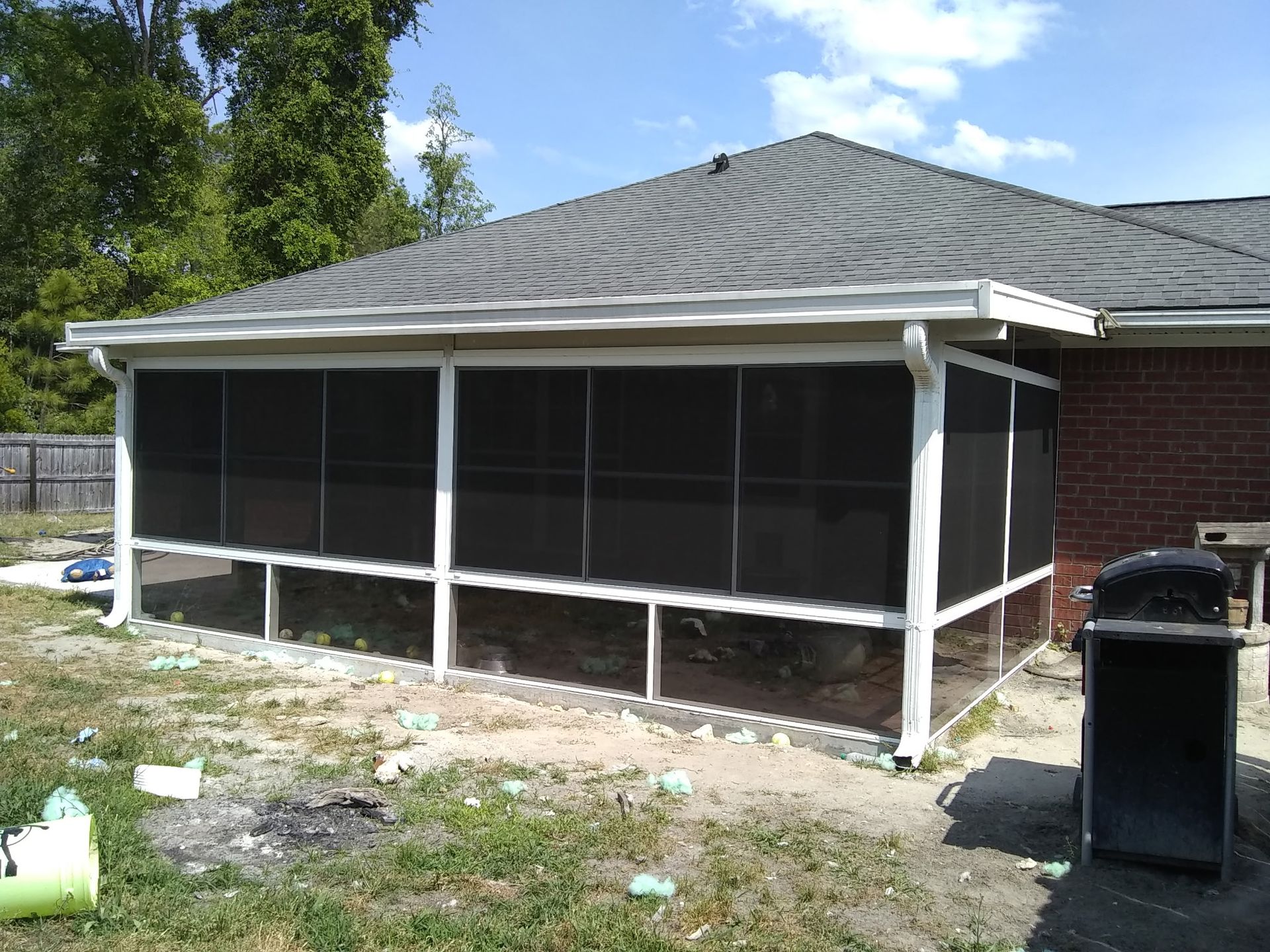 A white-framed screened-in porch attached to a brick house with a gray shingled roof, set in a grassy yard.