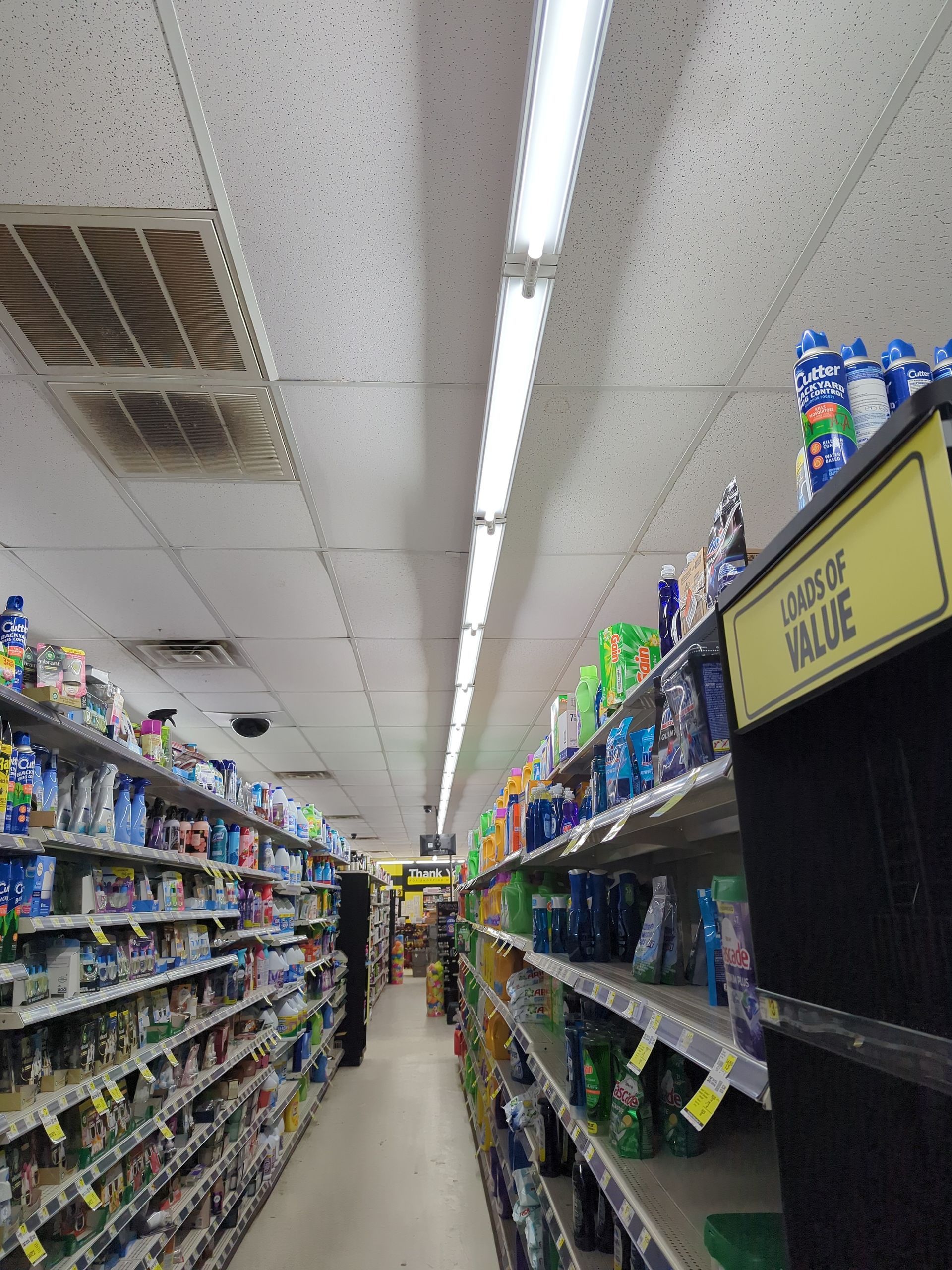 A long, brightly lit grocery aisle with shelves stocked with various cleaning and household products.
