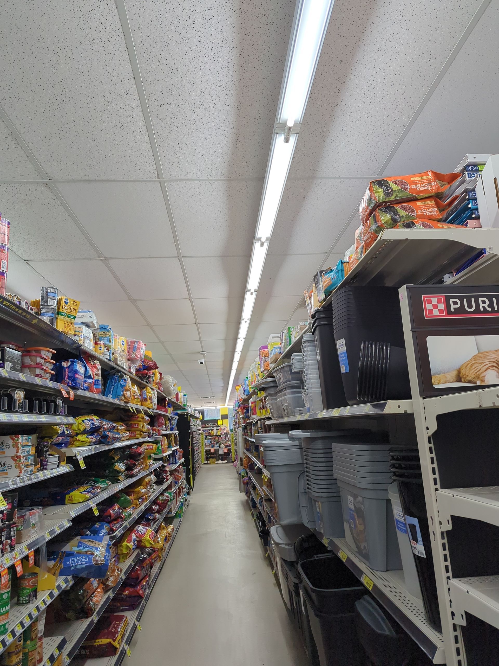 A narrow aisle in a grocery store with shelves stocked with various products on both sides and a bright overhead light.