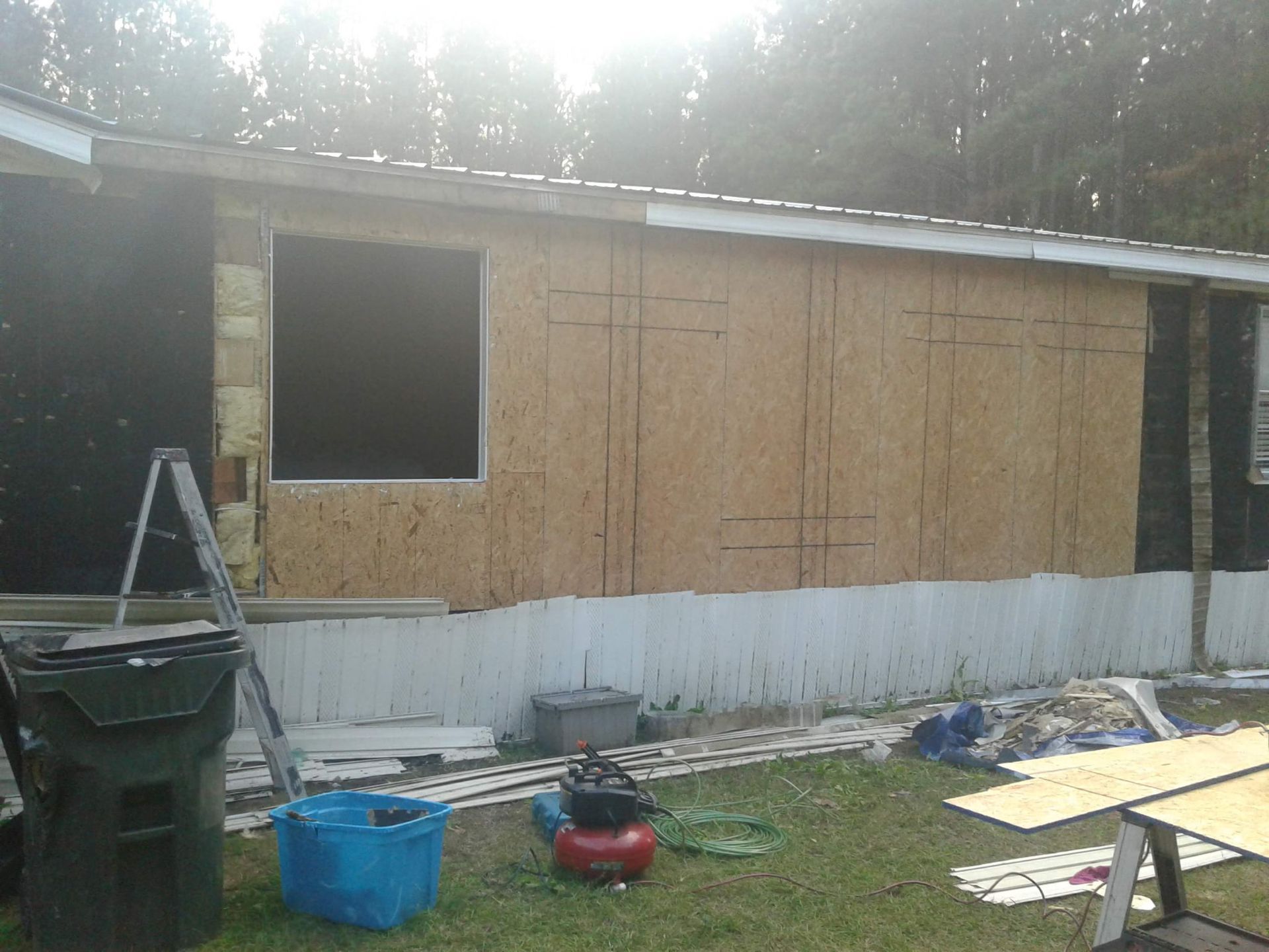 A mobile home exterior under renovation, featuring exposed plywood sheathing, a single window, and construction gear below.