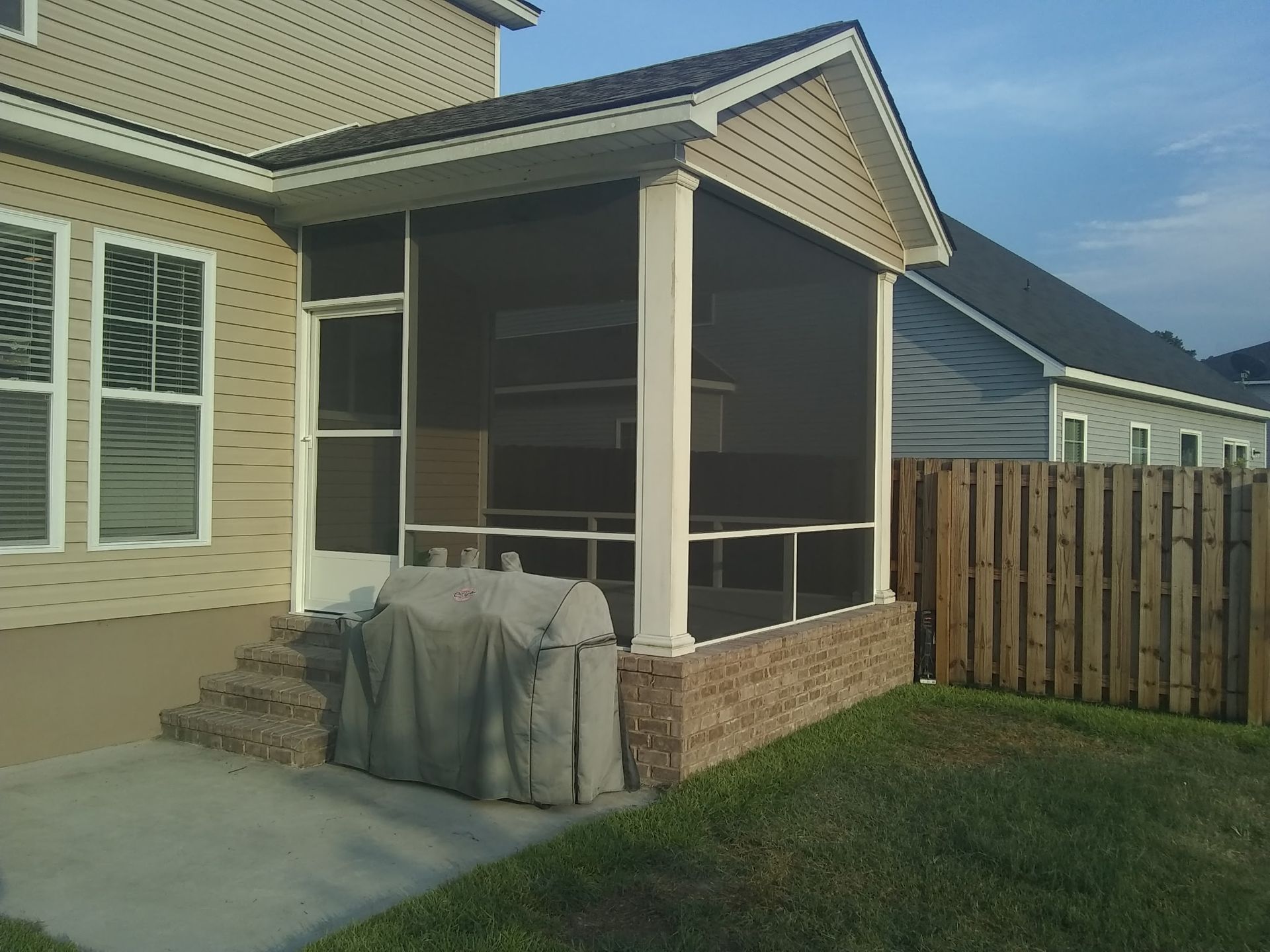 A beige house features a screened-in back porch with a brick base, a covered entry door, and a grill parked on the patio.