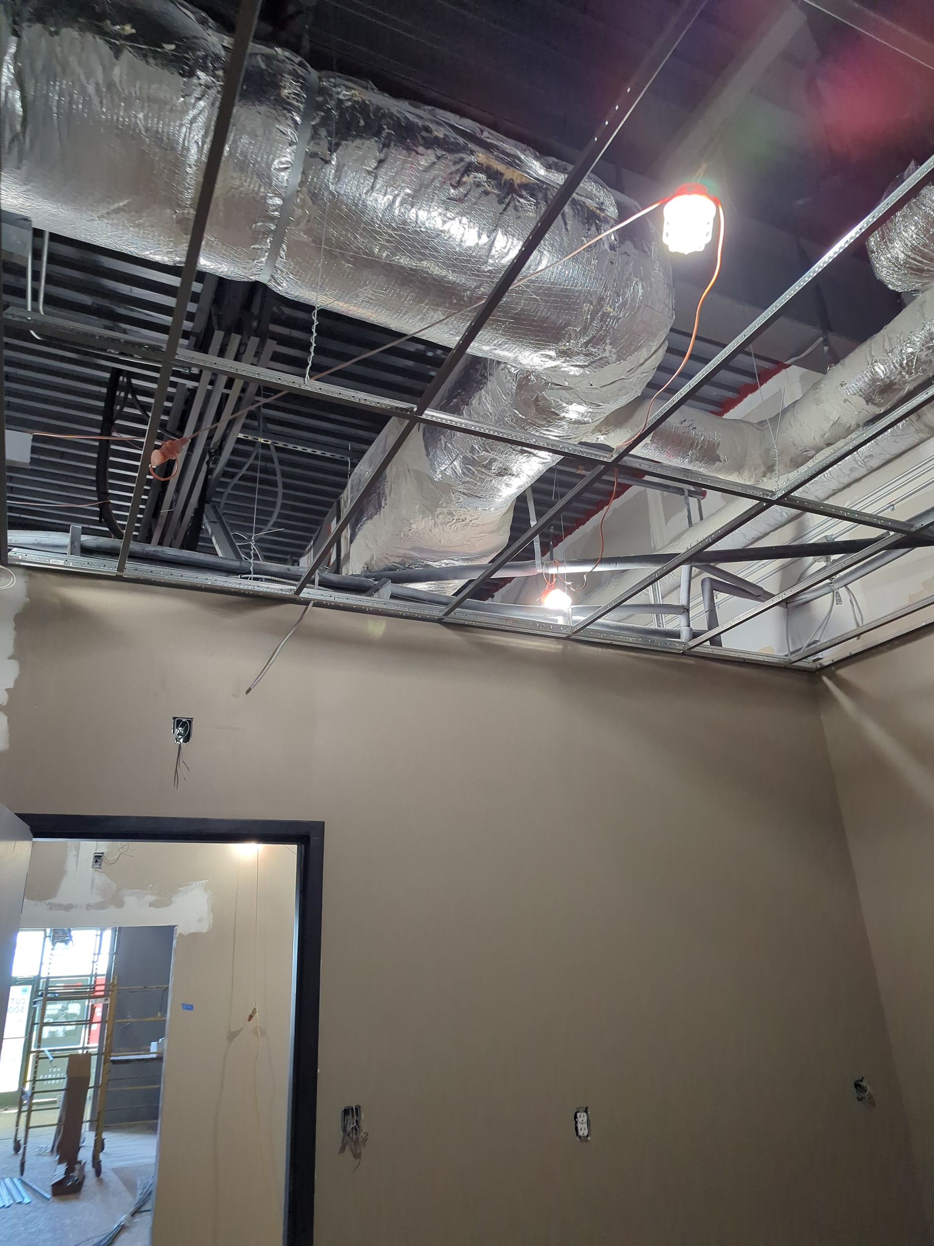 A room under construction with exposed ductwork, a metal ceiling grid, tan walls, and an open doorway.