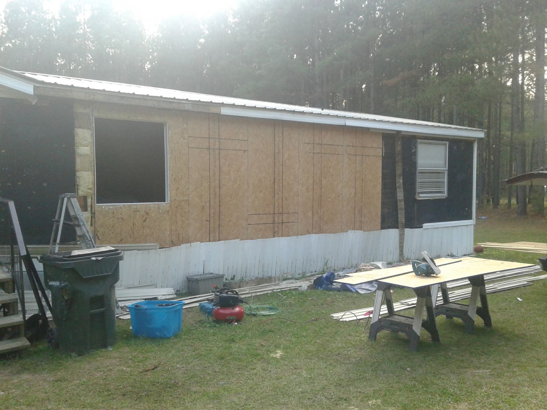 Exterior of a mobile home undergoing renovations with plywood sheathing on the side walls and a work table in the yard.