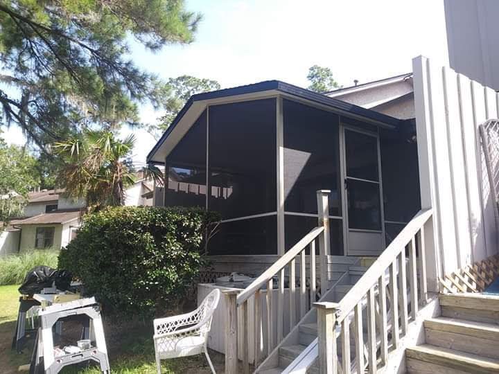 A screened-in porch with a white staircase, surrounded by trees and shrubs on a sunny day.