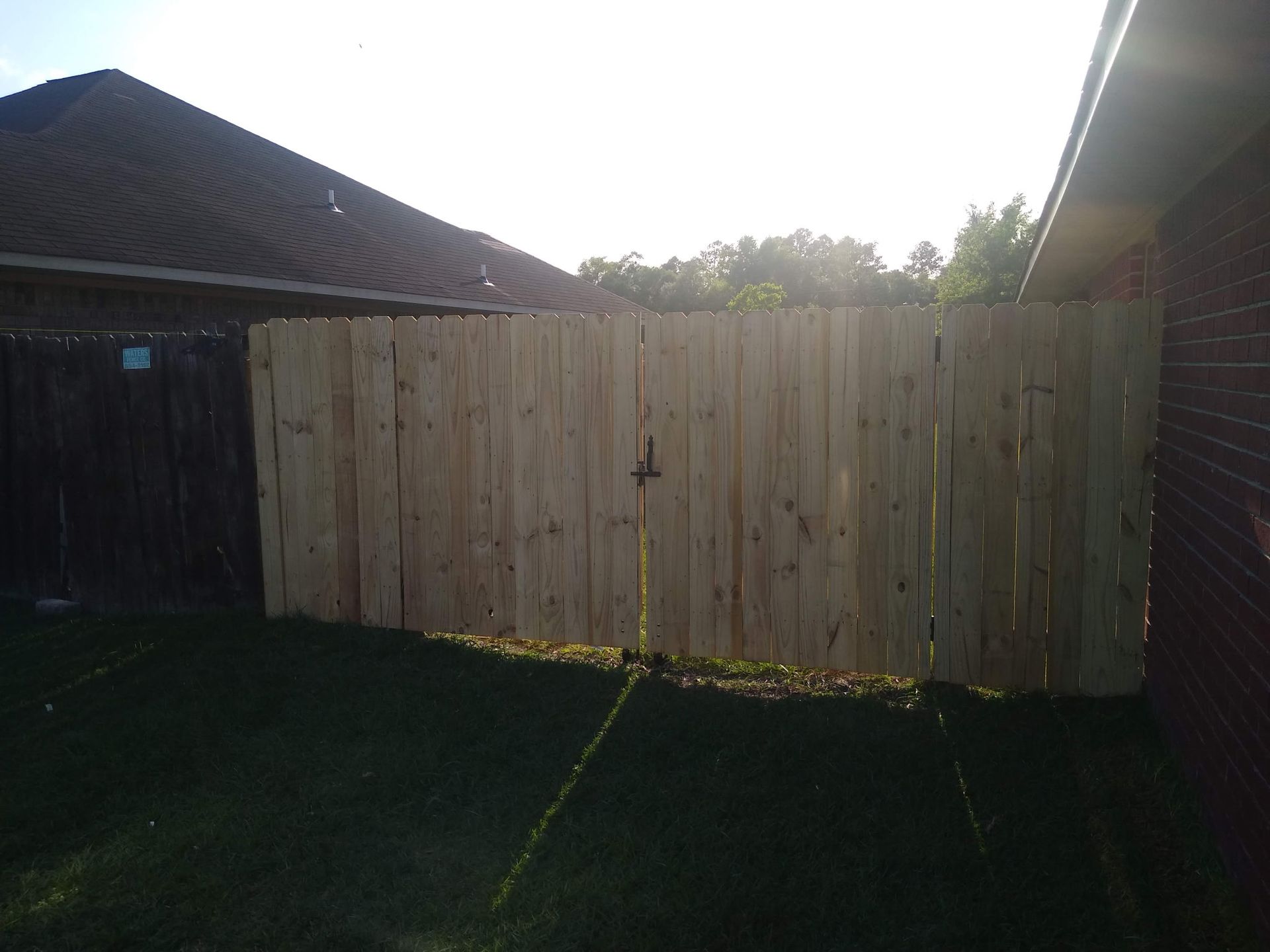 A new light-colored wooden fence sits between a brick house and a darker, weathered fence in a sunny backyard.