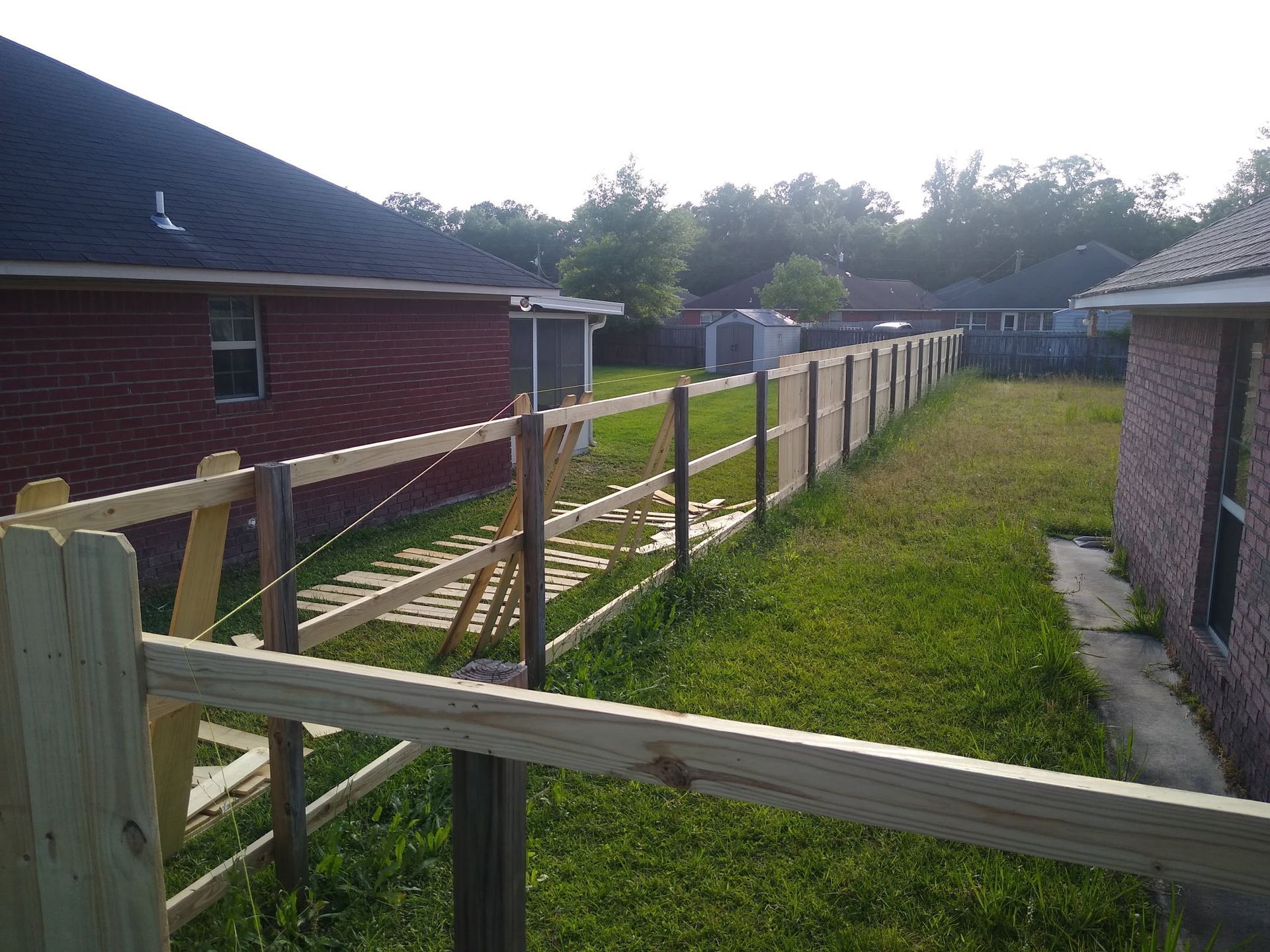 A partially constructed wooden fence runs between two brick houses in a grassy backyard.