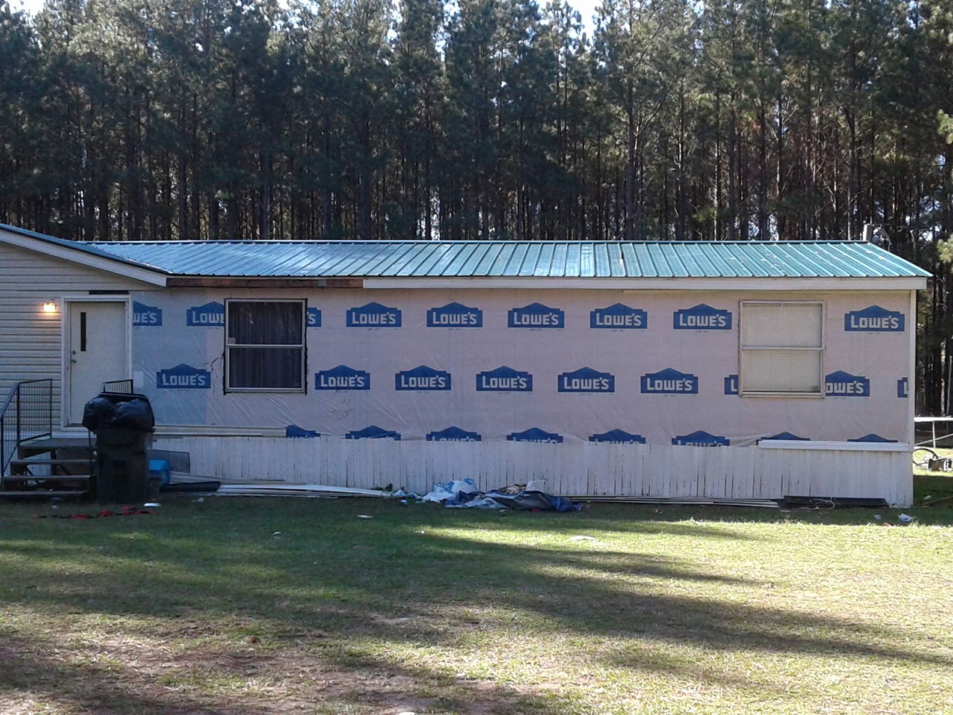 A side view of a mobile home exterior undergoing siding installation, featuring blue Lowe's house wrap and a green metal roof.