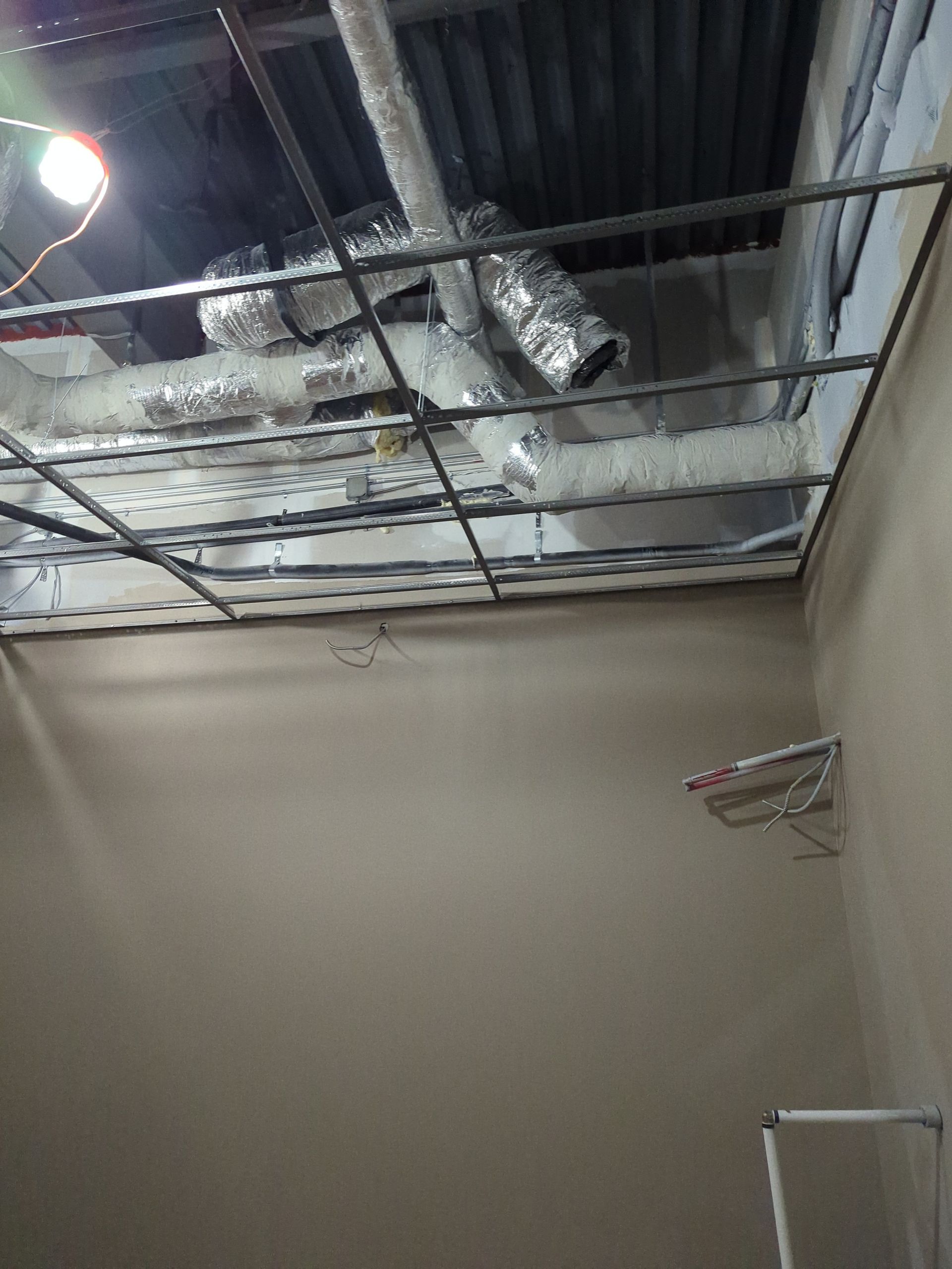 A view looking up at exposed ceiling infrastructure with ductwork and a metal grid frame in a room with beige walls.