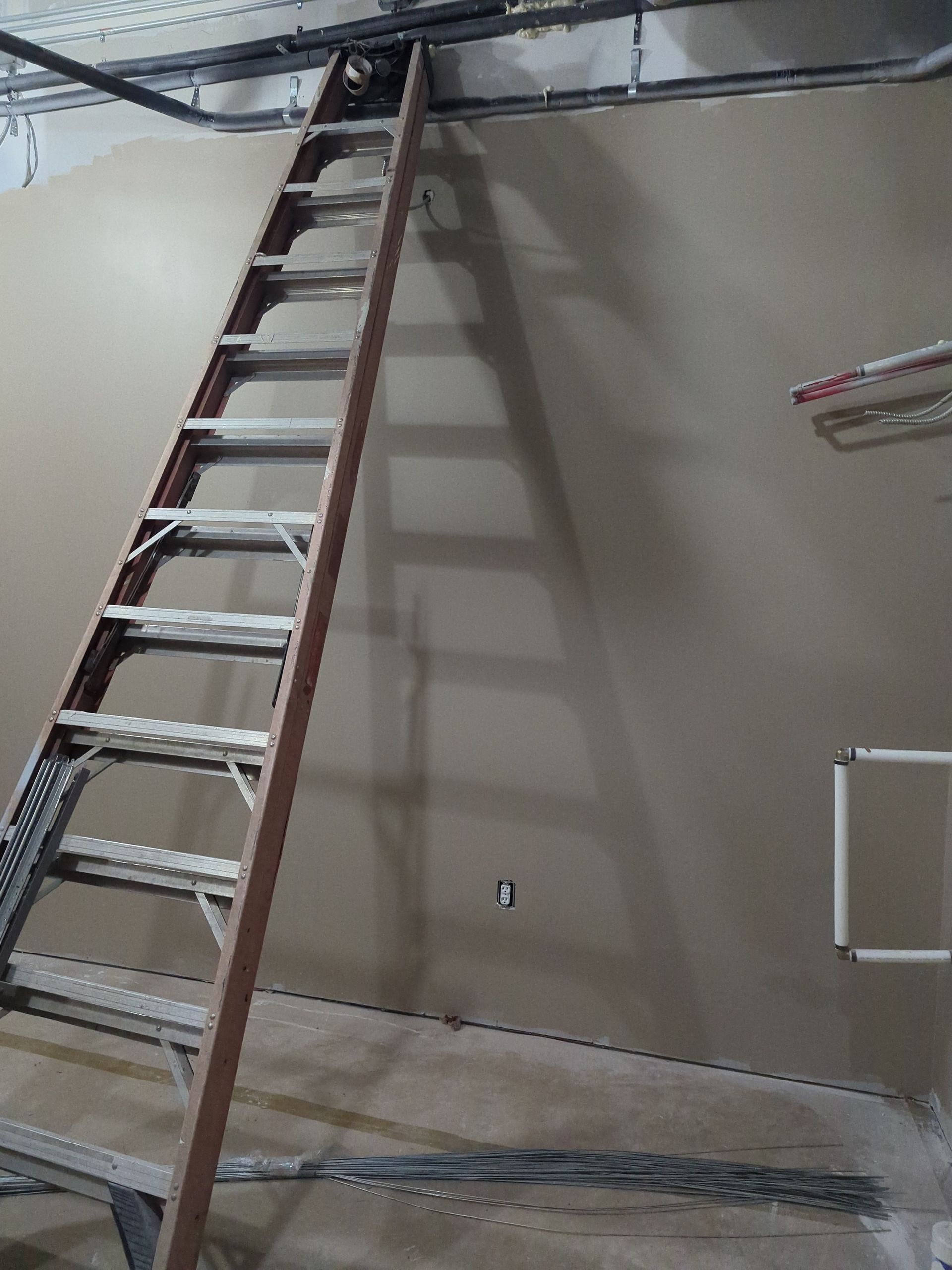 An extension ladder leans against a finished drywall partition in a room under construction with exposed overhead pipes.