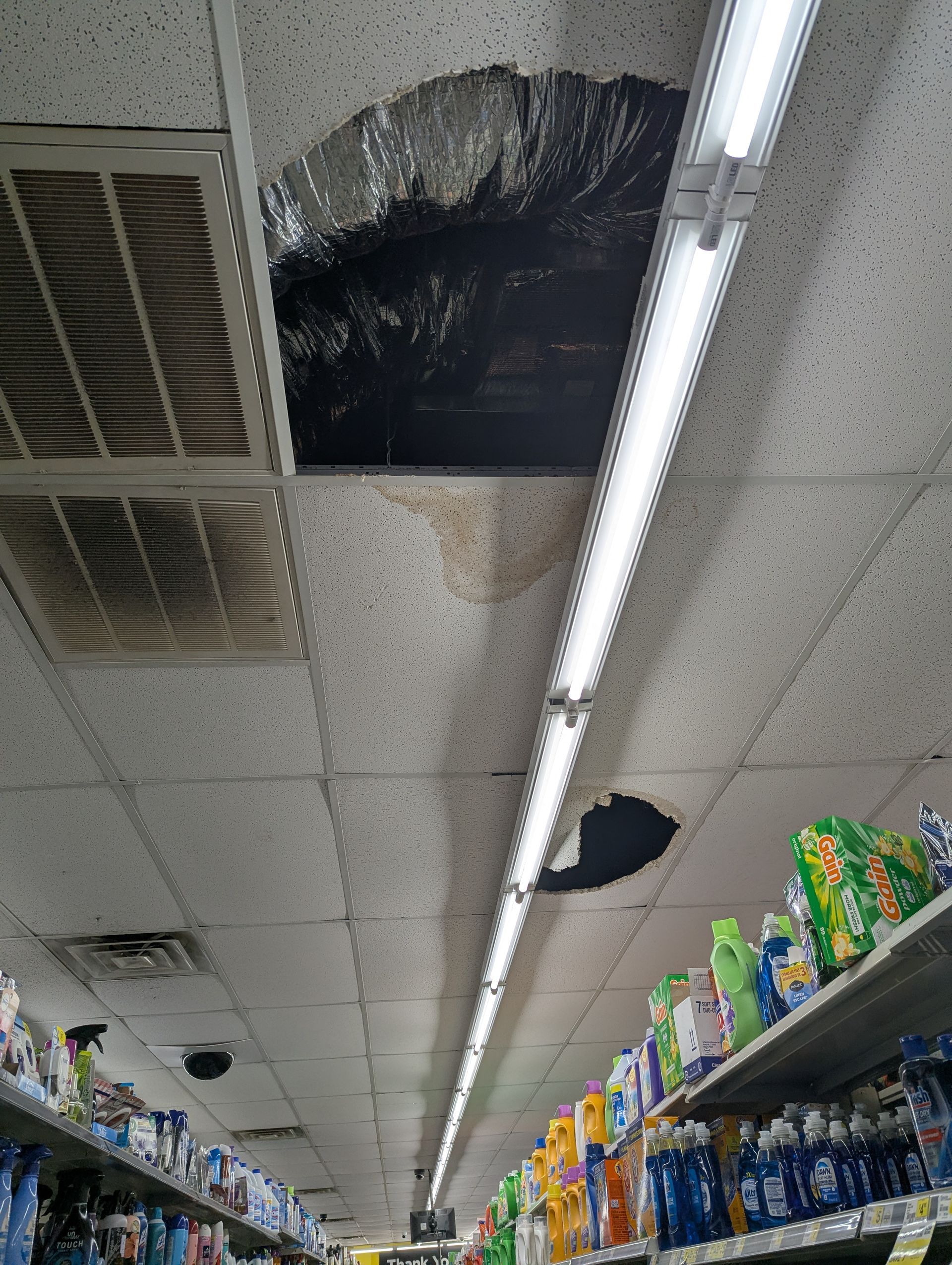 A damaged drop ceiling in a store aisle with two large holes and water stains near a long fluorescent light fixture.