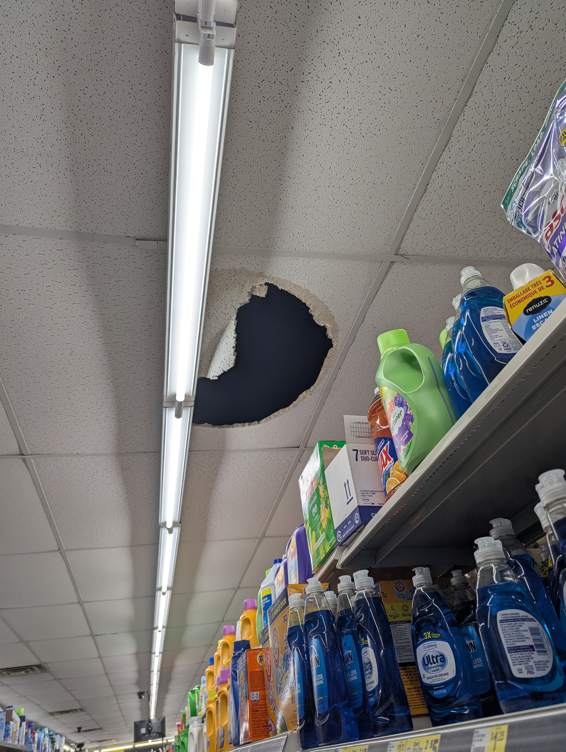 A large hole in the drop ceiling of a retail store aisle, located next to a long fluorescent light fixture.