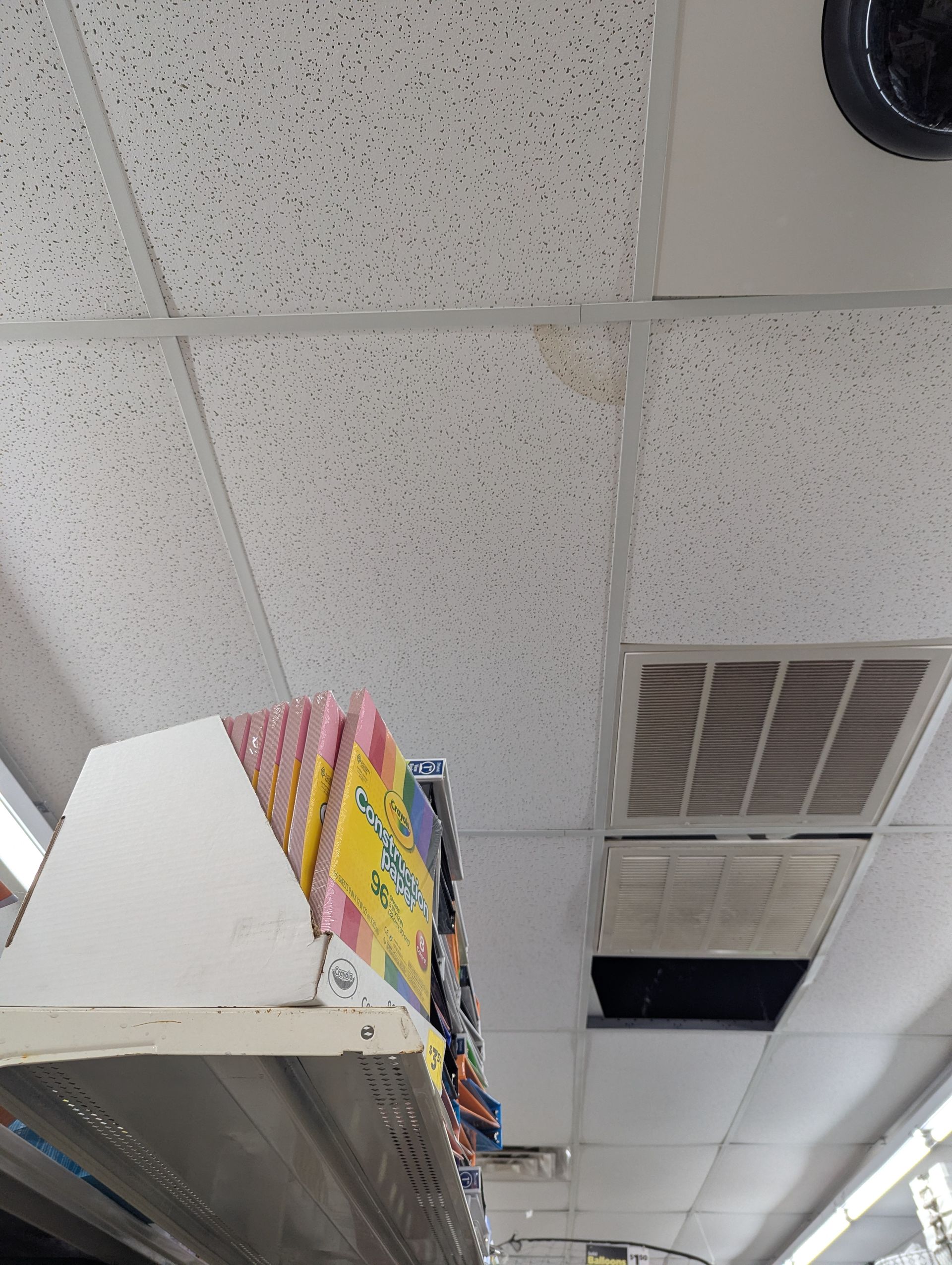 A retail shelf corner shown from a low angle, highlighting a water-stained, damaged ceiling tile and a missing vent cover.