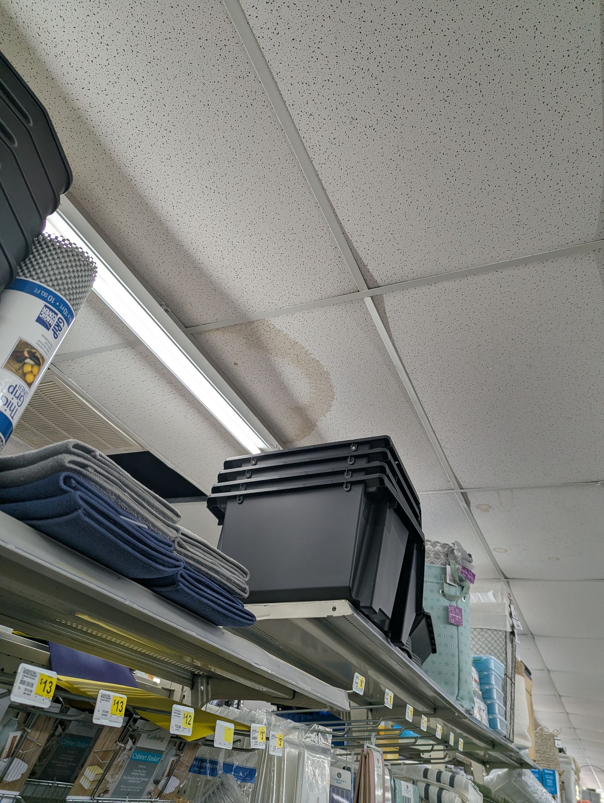 A view looking up at a store shelf holding a stack of black plastic bins and folded gray fabric near a stained ceiling.