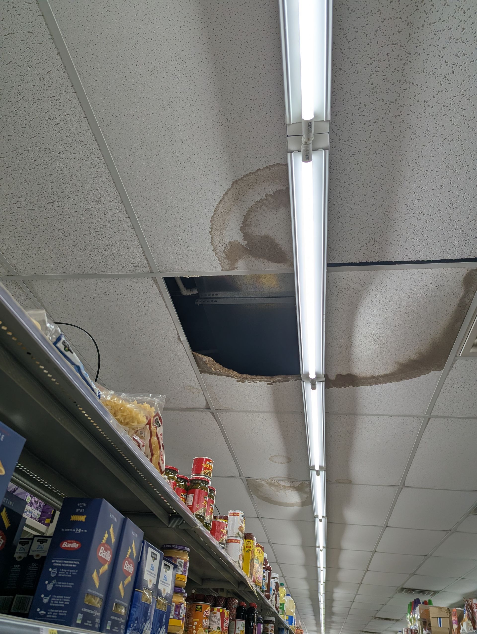 A grocery store aisle with a water-damaged, crumbling ceiling tile and a missing tile near a long overhead fluorescent light.