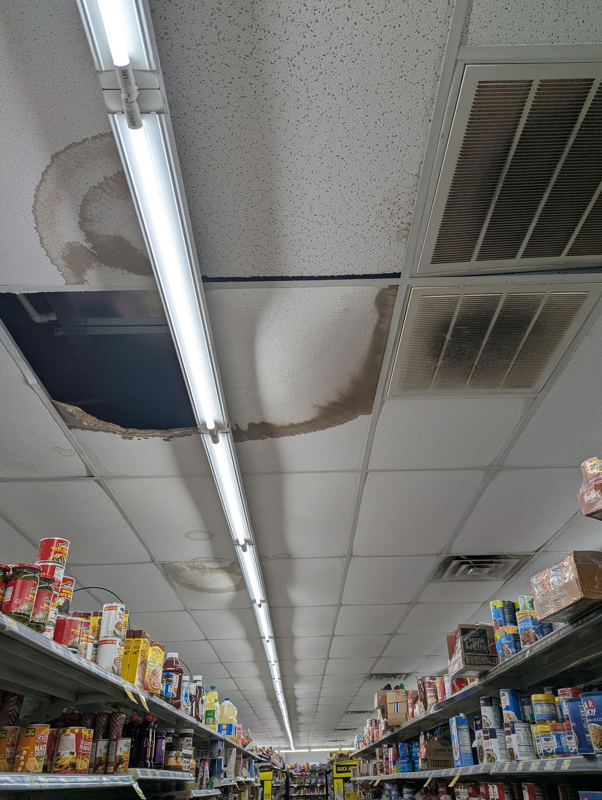 A retail aisle with damaged drop ceiling tiles, including a missing tile and large water stains next to an air vent.