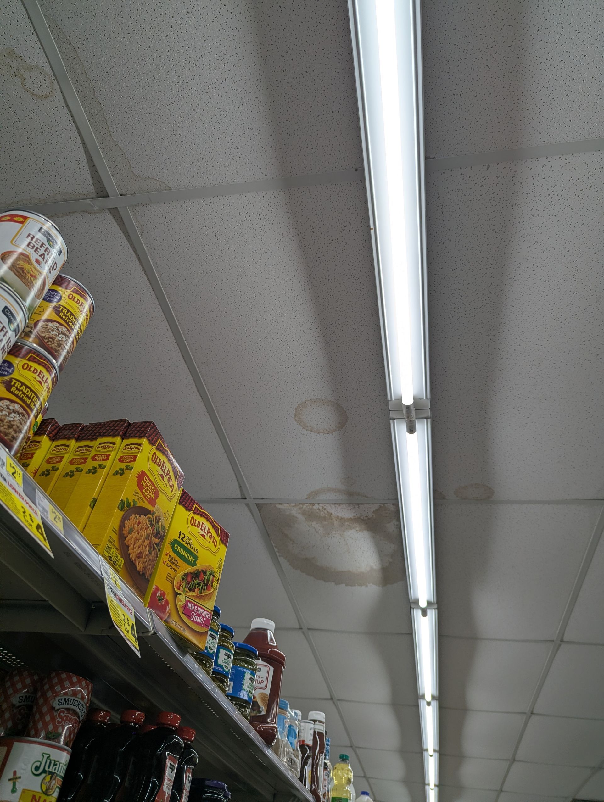 A store ceiling shows water damage stains next to a long, glowing fluorescent light fixture above retail shelves.