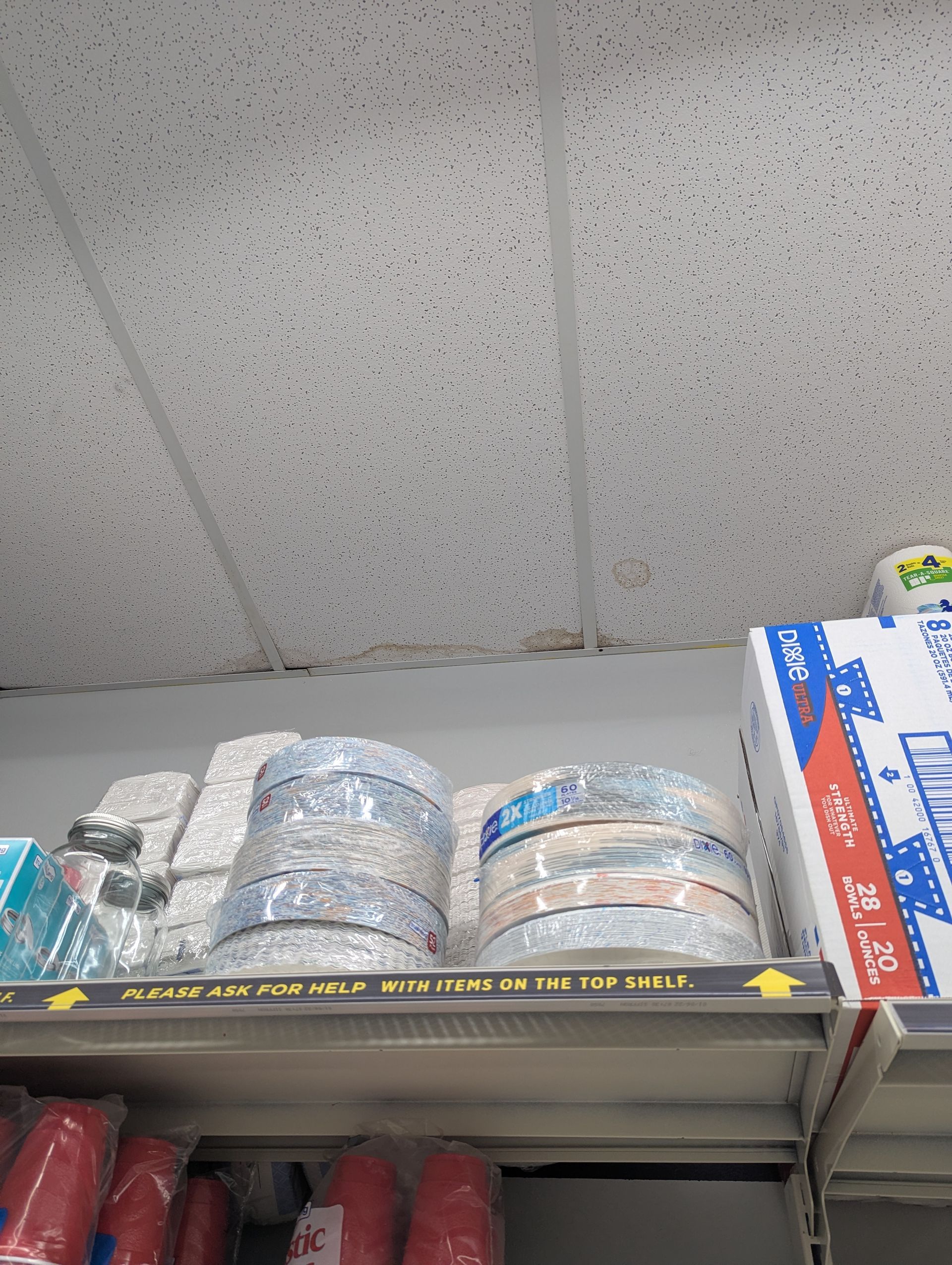 Retail shelves stocked with rolls of ribbon, plastic cups, and boxes of paper goods near a drop ceiling.