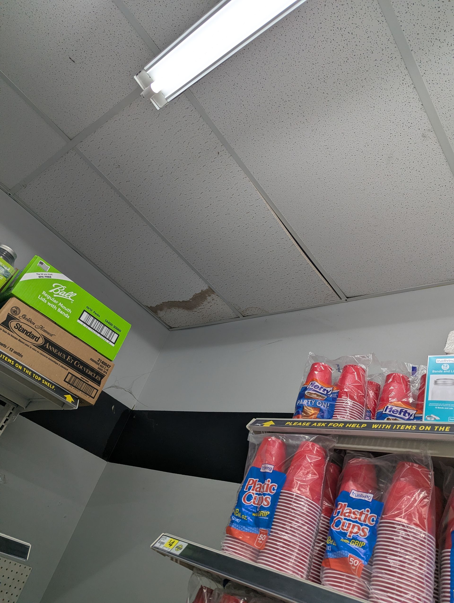 A retail shelf displaying stacked red plastic cups beneath a stained, damaged ceiling tile in a store.