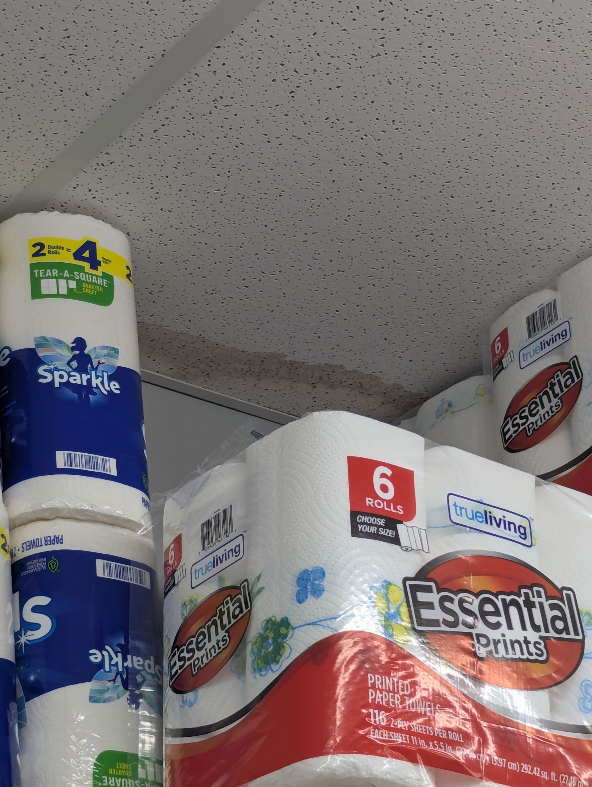 Packages of Sparkle and Brawny paper towels stacked on a retail shelf against a drop ceiling.