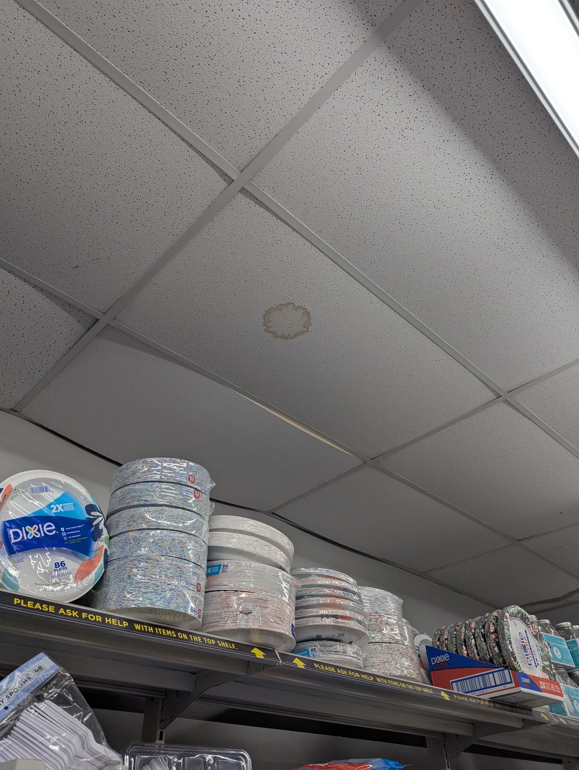 Stacks of paper plates and bowls sit on a shelf below a drop ceiling with a water stain.