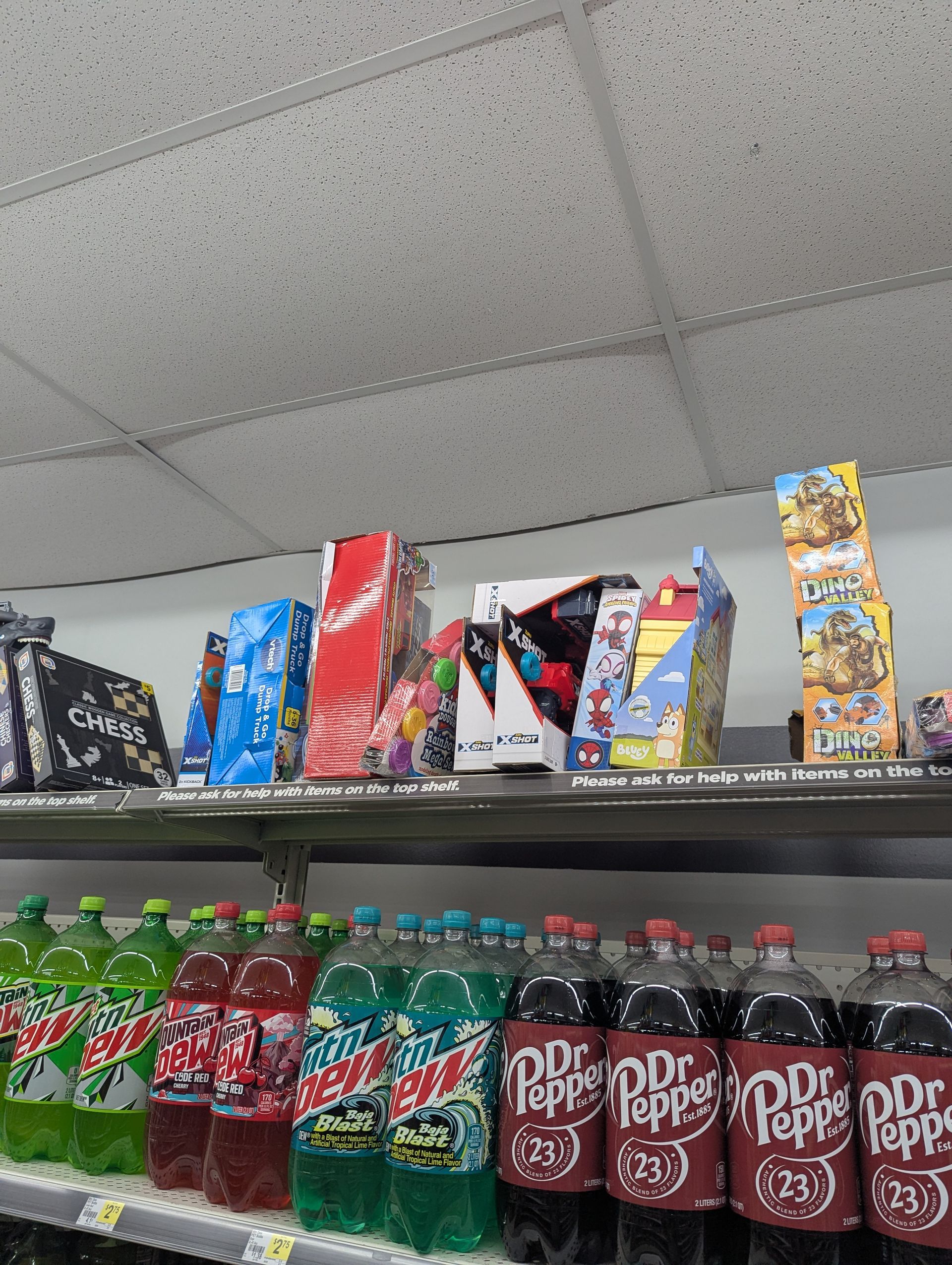 Store shelf with rows of soda bottles, including Mountain Dew and Dr. Pepper, and miscellaneous snack boxes on the top shelf.