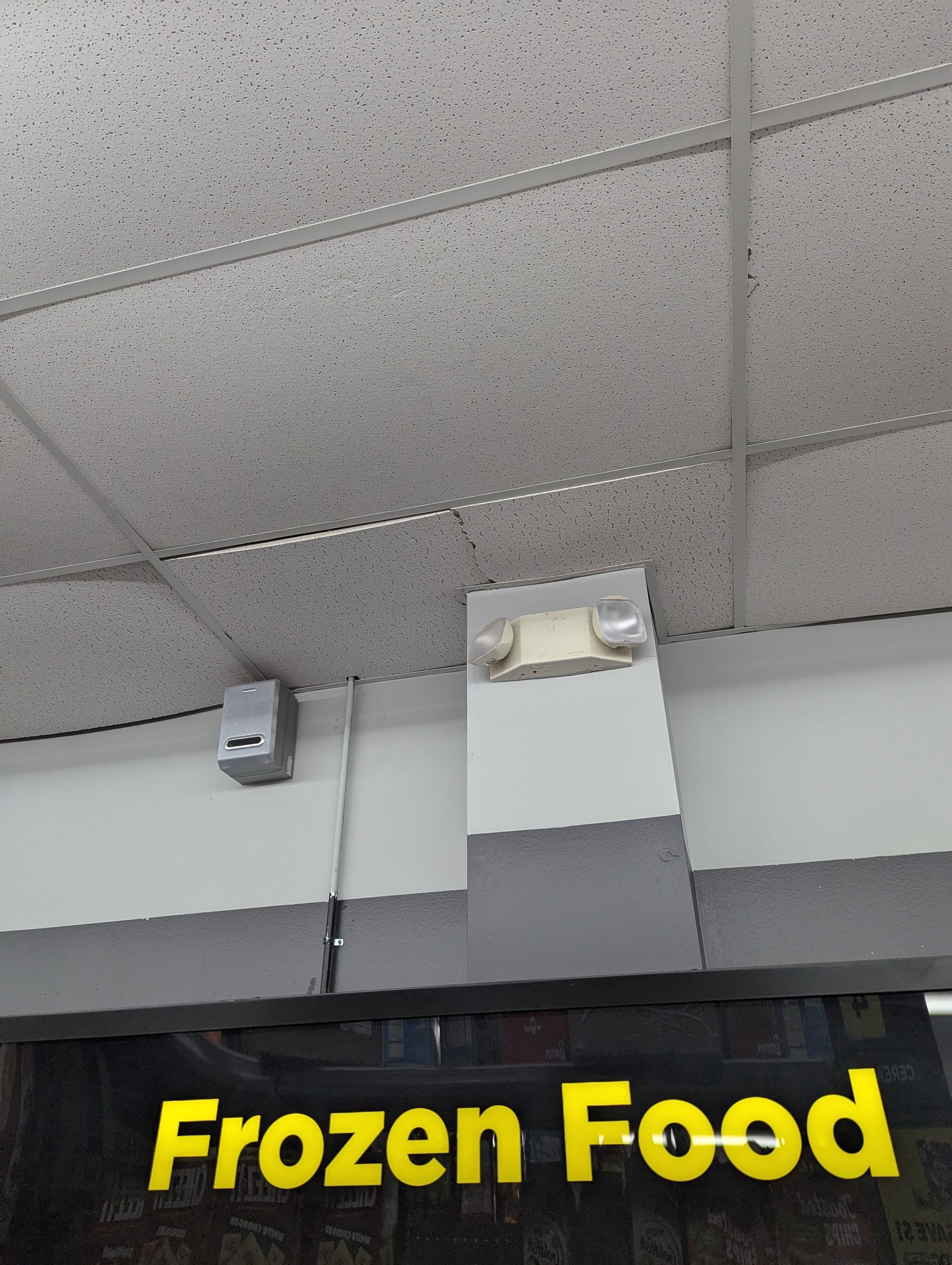 An emergency light and a small grey sensor mounted on a wall above a black sign that reads 