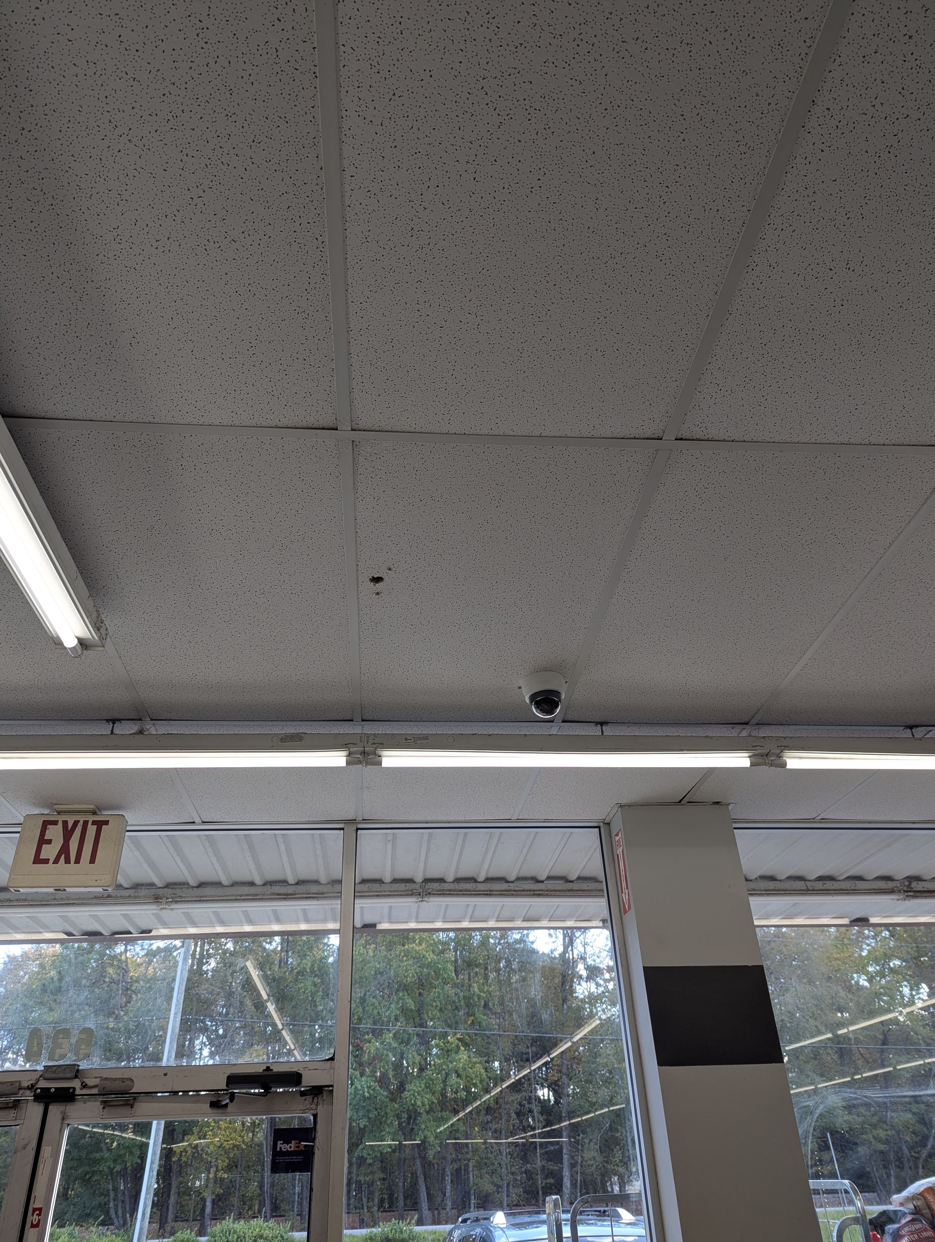 A security camera mounted on a textured white ceiling above a glass store entrance and an exit sign.