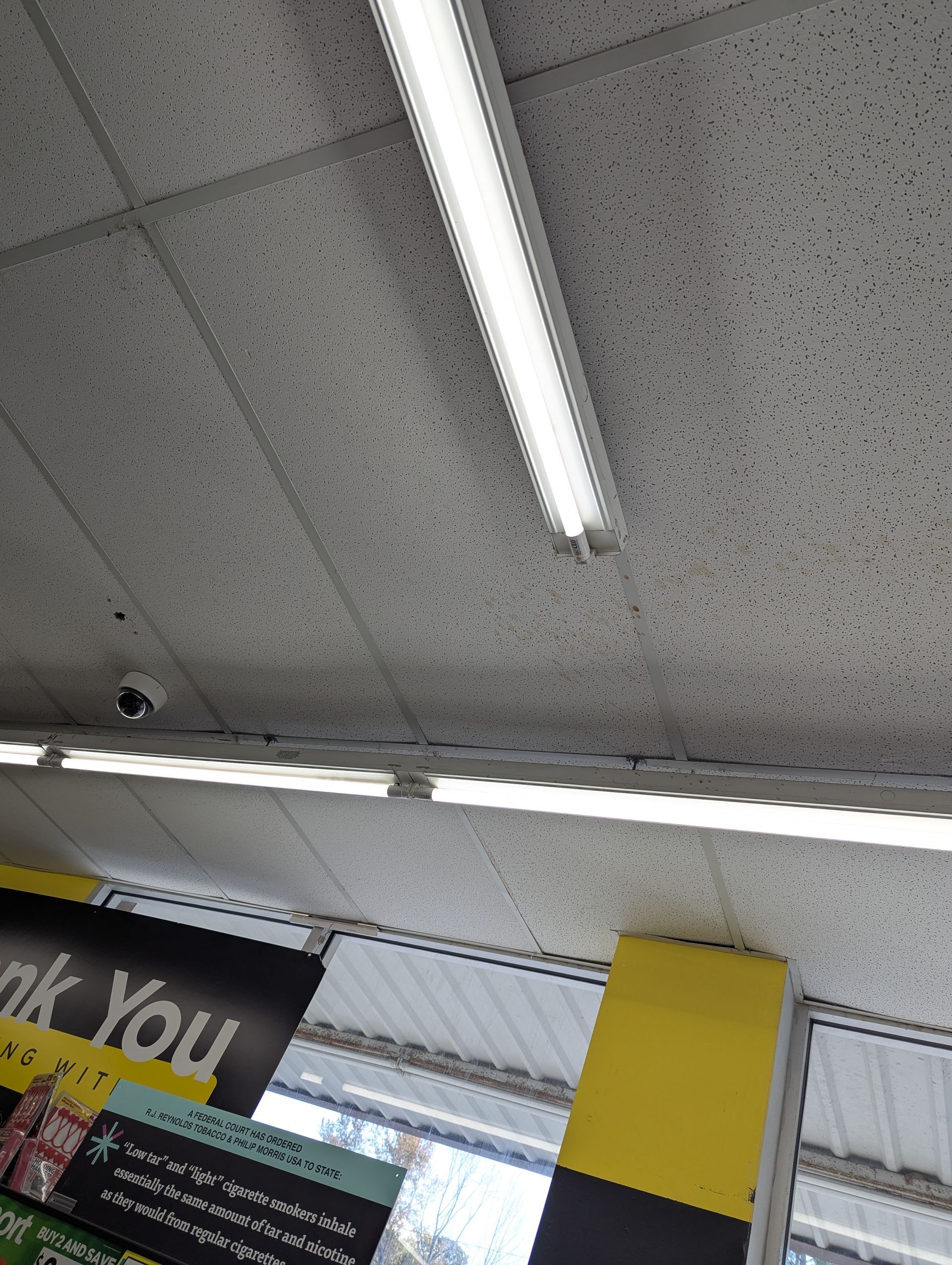 A drop ceiling with fluorescent lights and a security camera, featuring a partially visible sign with yellow accents.
