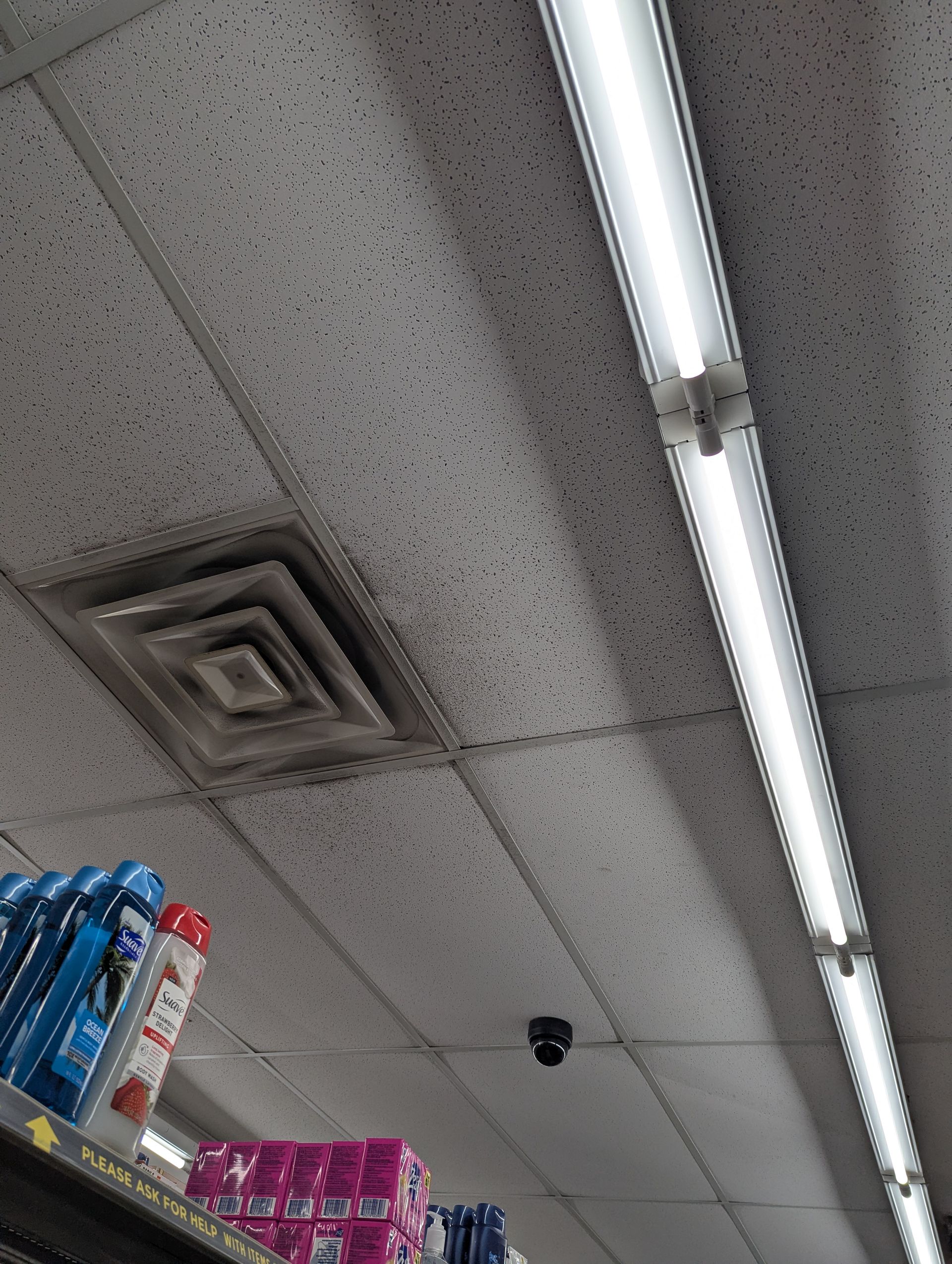 A view of a commercial drop ceiling showing a dirty HVAC vent, a security camera, and a long fluorescent light fixture.