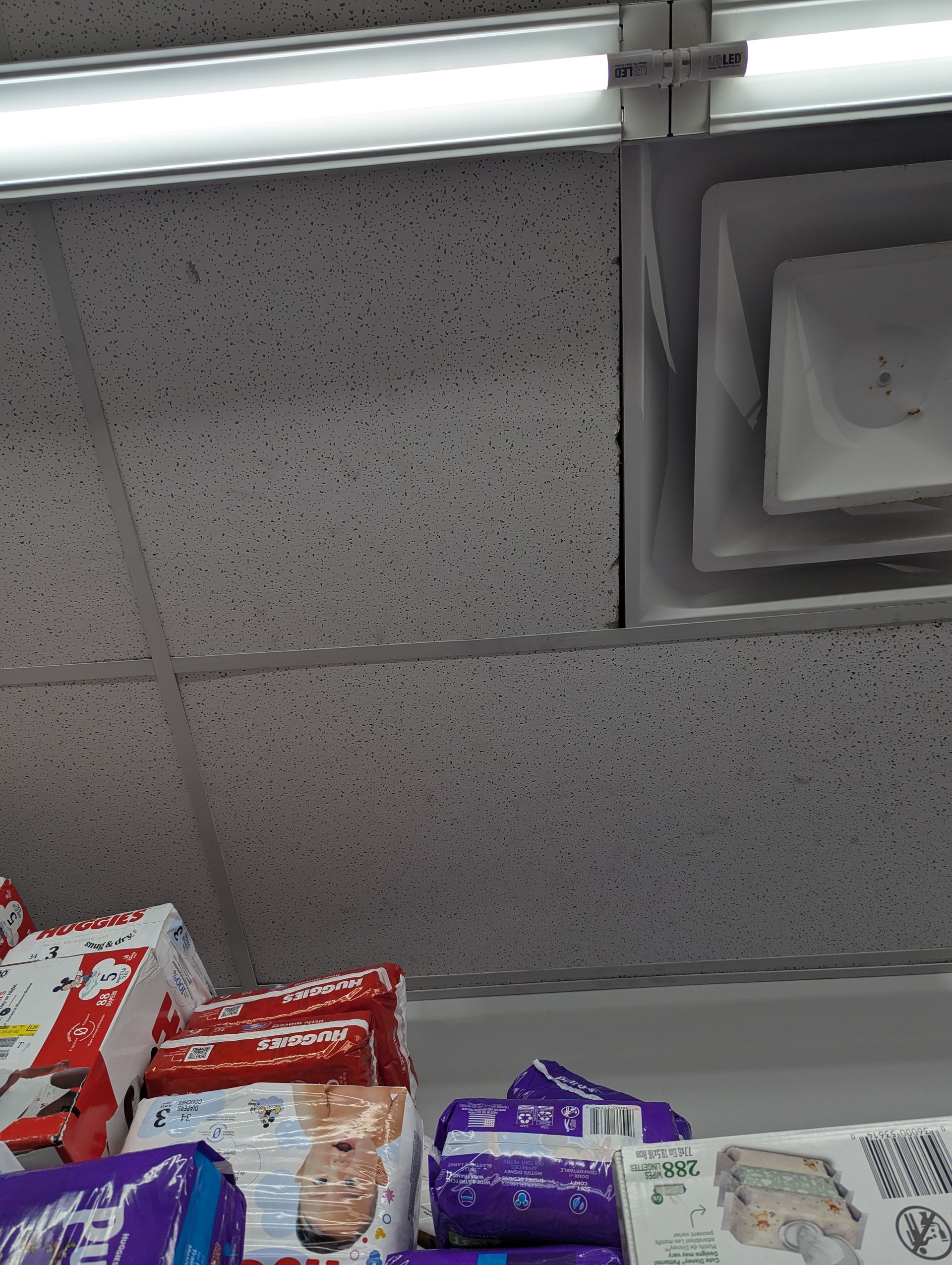 A low-angle view of retail shelves filled with various brands of diapers below a ceiling with an air vent and lights.