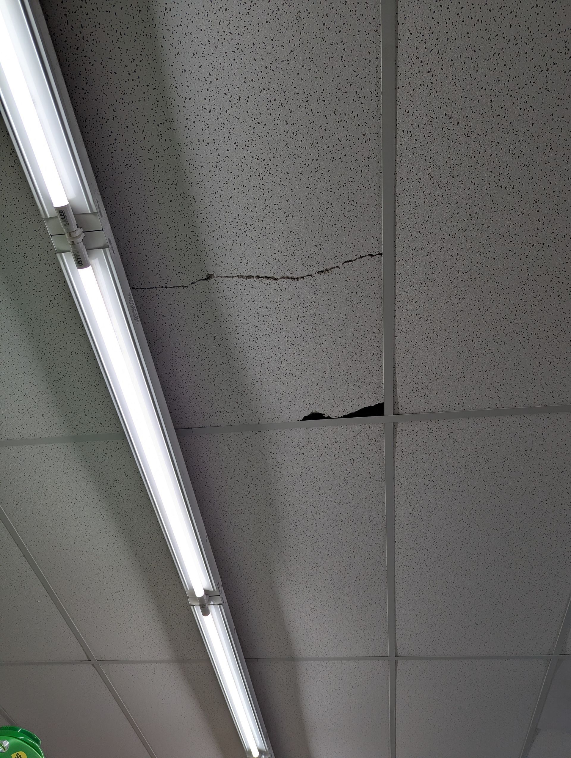 A cracked and damaged drop ceiling tile next to an overhead fluorescent light fixture.
